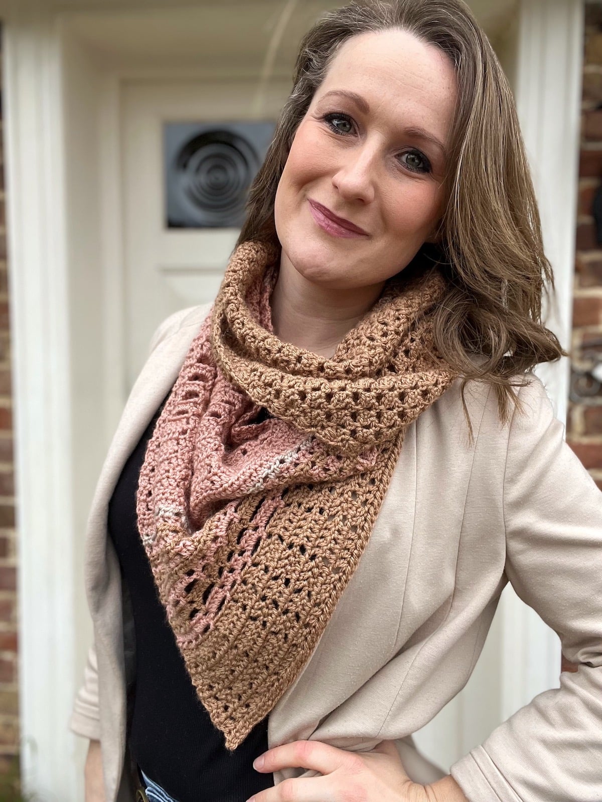 Woman standing outside in front of a door, wearing a beige jacket, black shirt, and a brown and pink scarf made from a lightweight crochet scarf pattern, looking at the camera with a slight smile.
