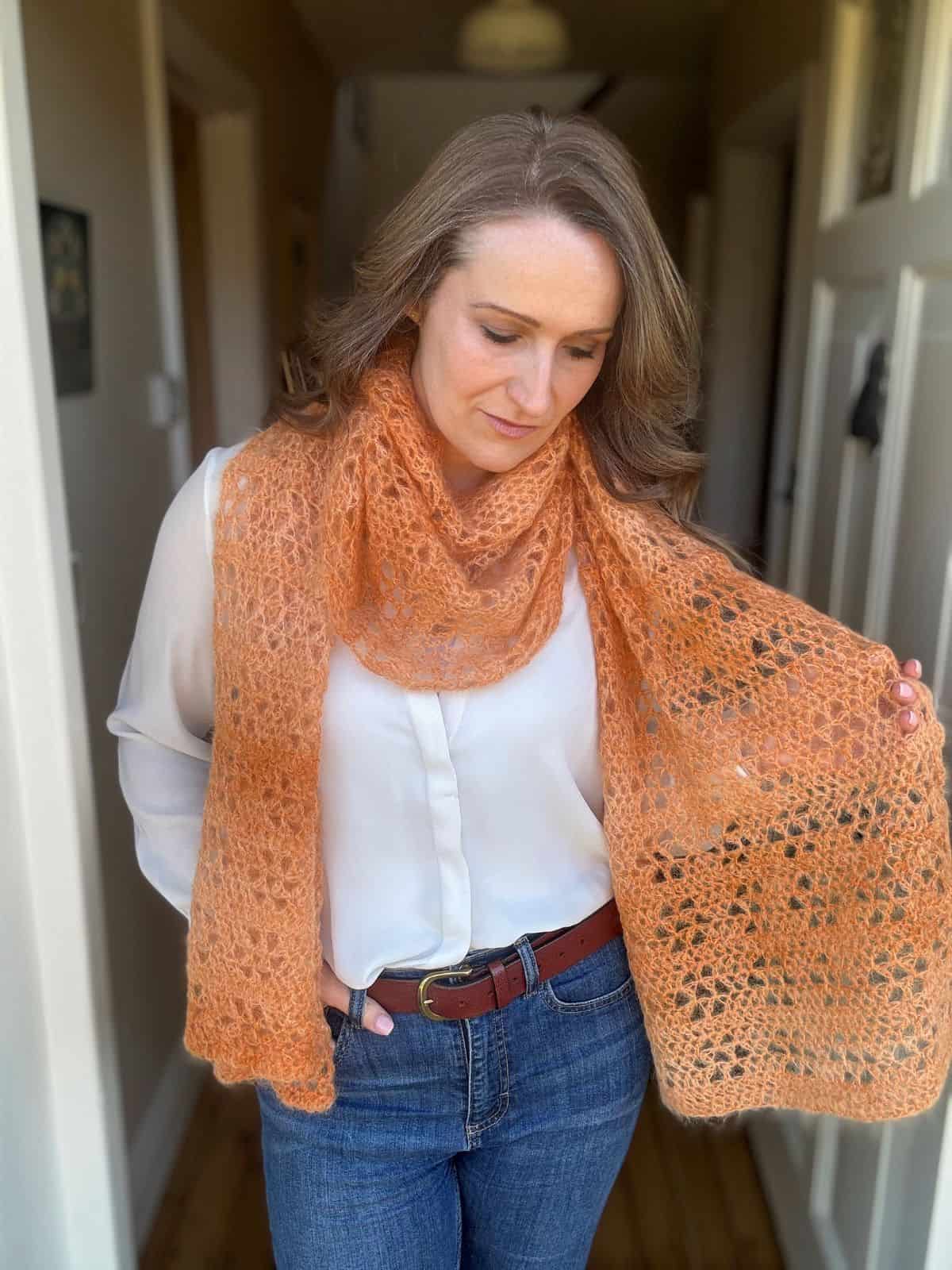 A person stands in a hallway wearing a light blouse and jeans, accentuated by an elegant orange crocheted scarf draped around their neck and over one shoulder.