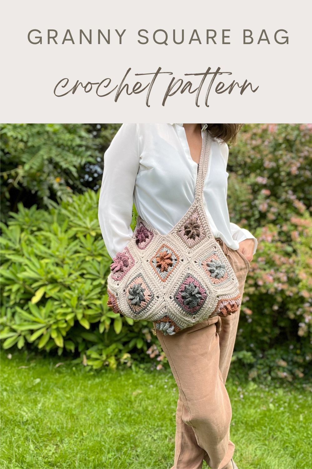 A person stands outdoors in a white shirt and tan pants, carrying a stylish granny square bag crochet over their shoulder. Shrubs and grass add a natural touch to the background.