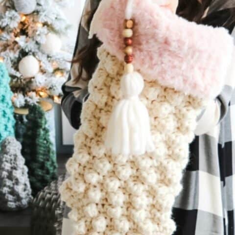 A woman is holding a crocheted sock in front of a christmas tree.