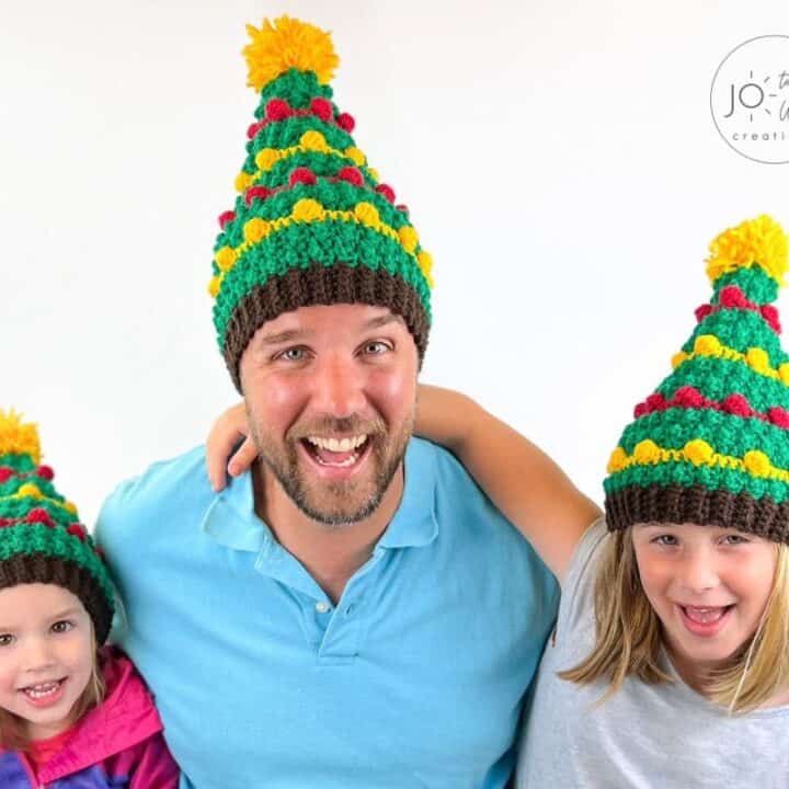 A man and his two daughters are posing for a photo in a knitted christmas tree hat.