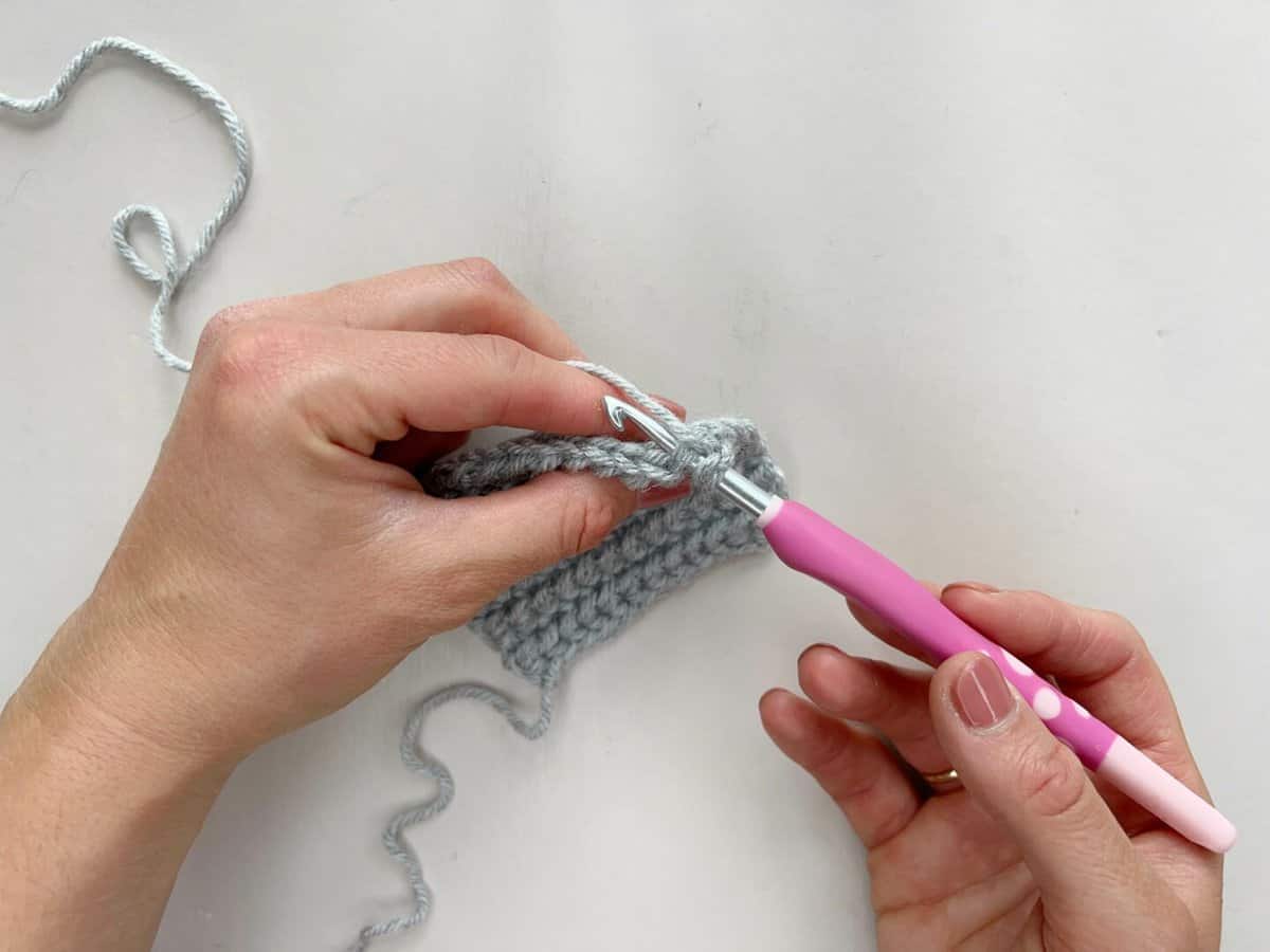 A person using a pink crochet hook to insert the hook to make a yarn over slip stitch.