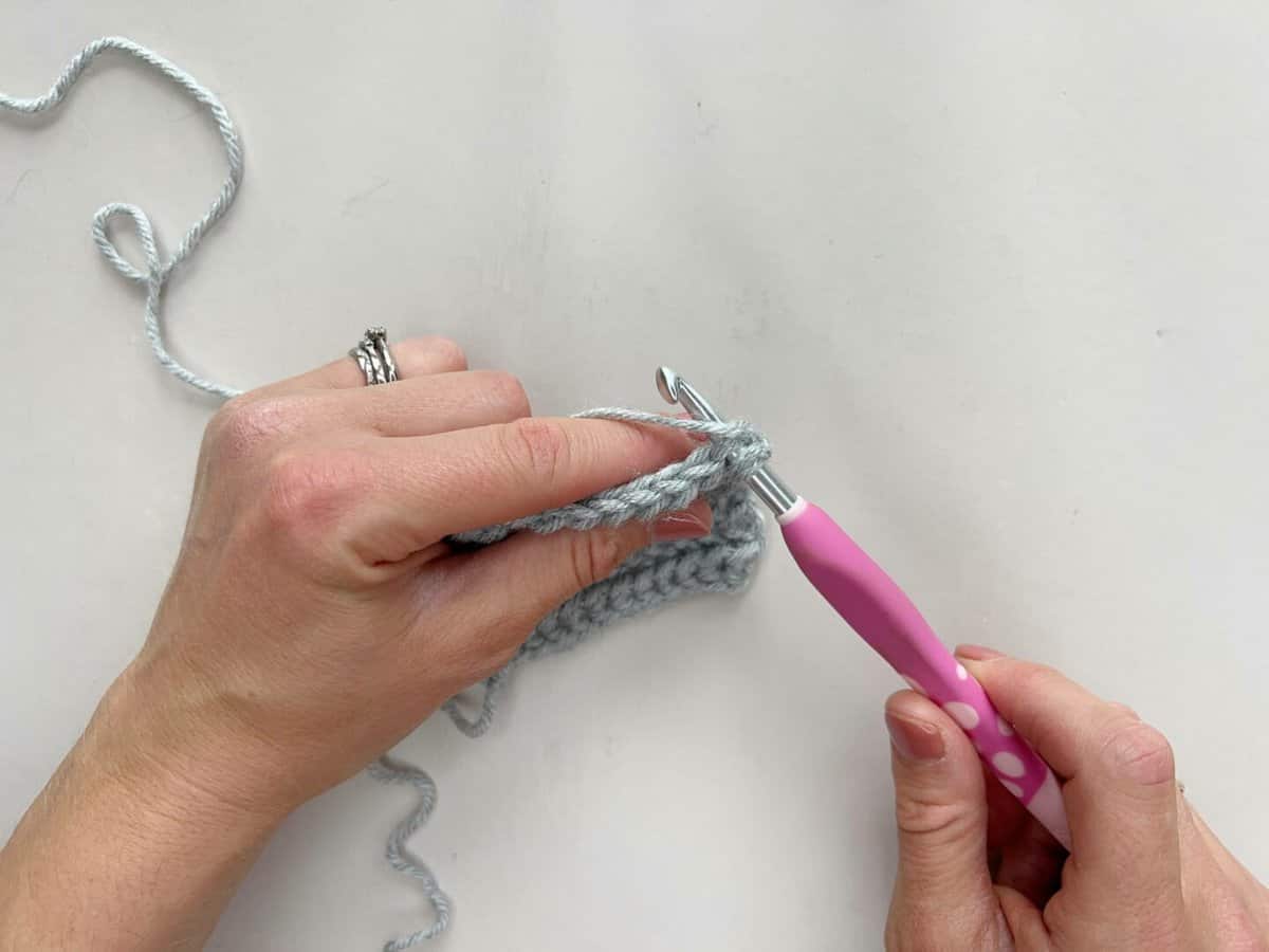 A person is using a pink crochet hook to yarn over and make a crochet yarn over slip stitch.