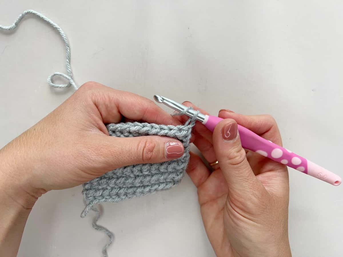 A person is using a pink crochet hook to yarn over and start to crochet the yarn over slip stitch.