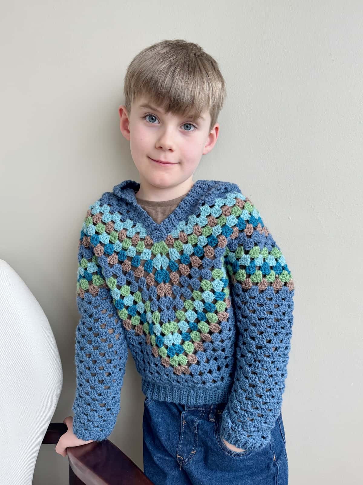 A young boy wearing a child crochet hoodie leaning against a chair.