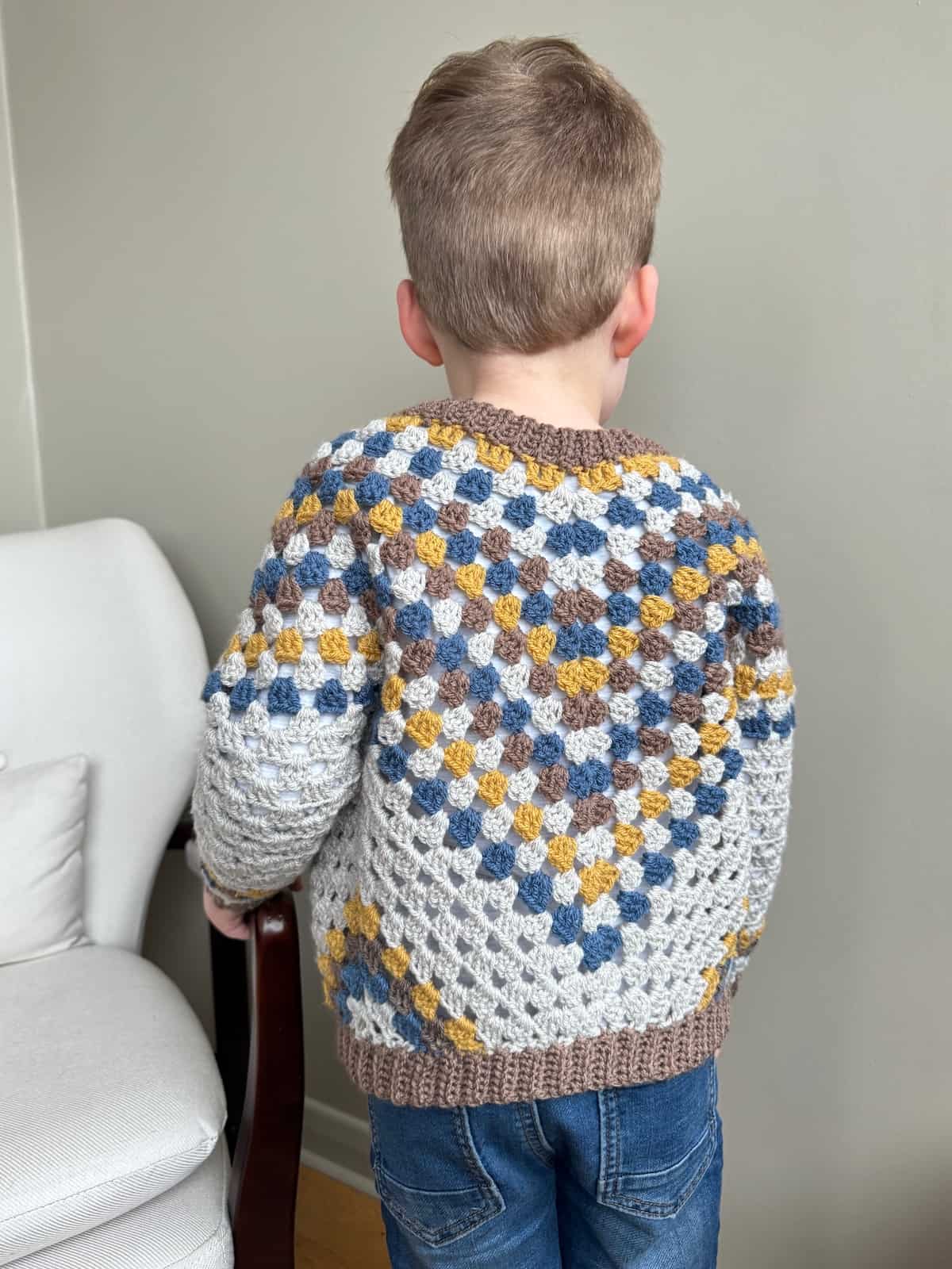 A young boy wearing a colourful crochet cardigan standing in front of a chair.