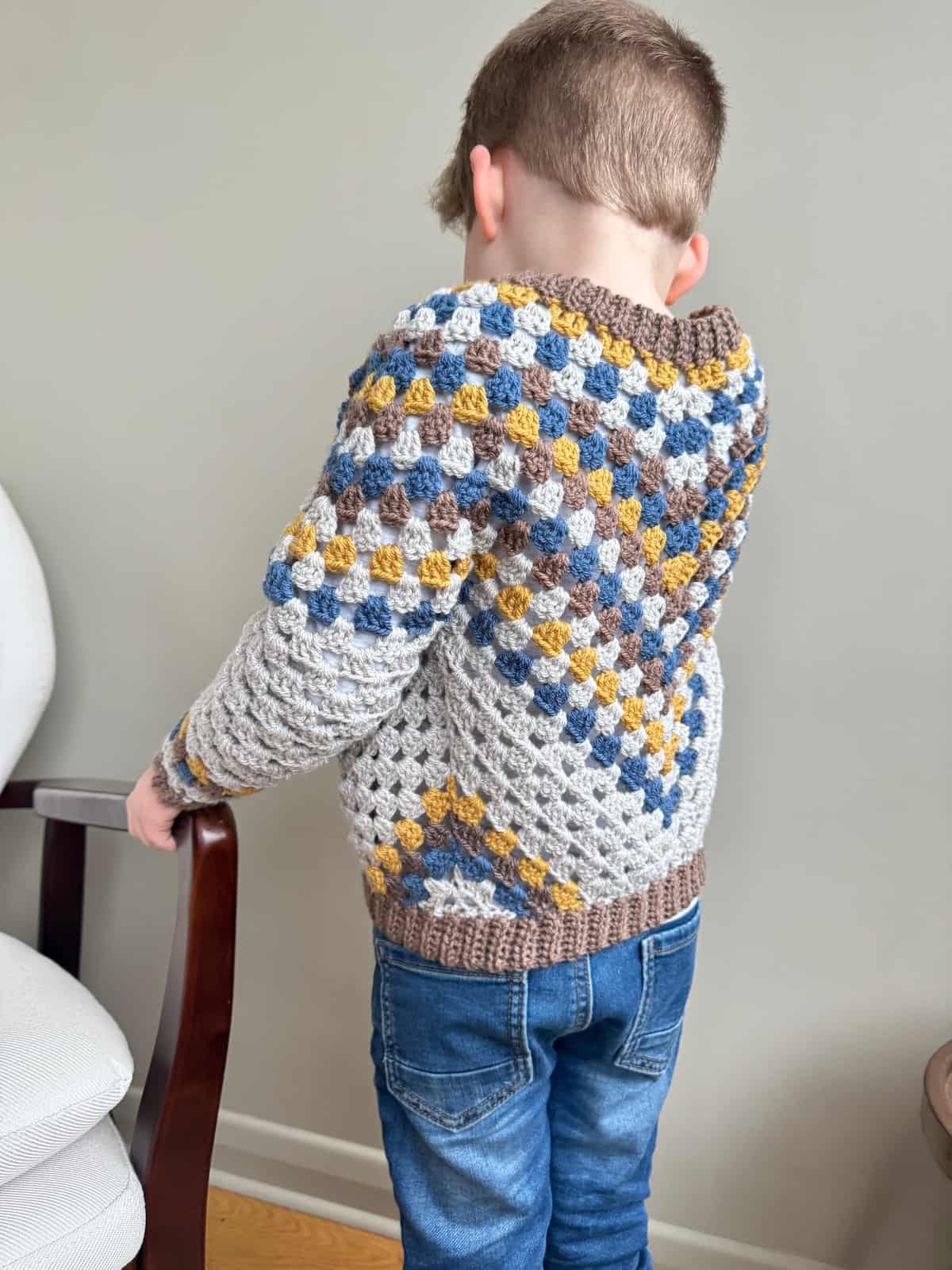 A young boy wearing a crochet cardigan stands in front of a chair.