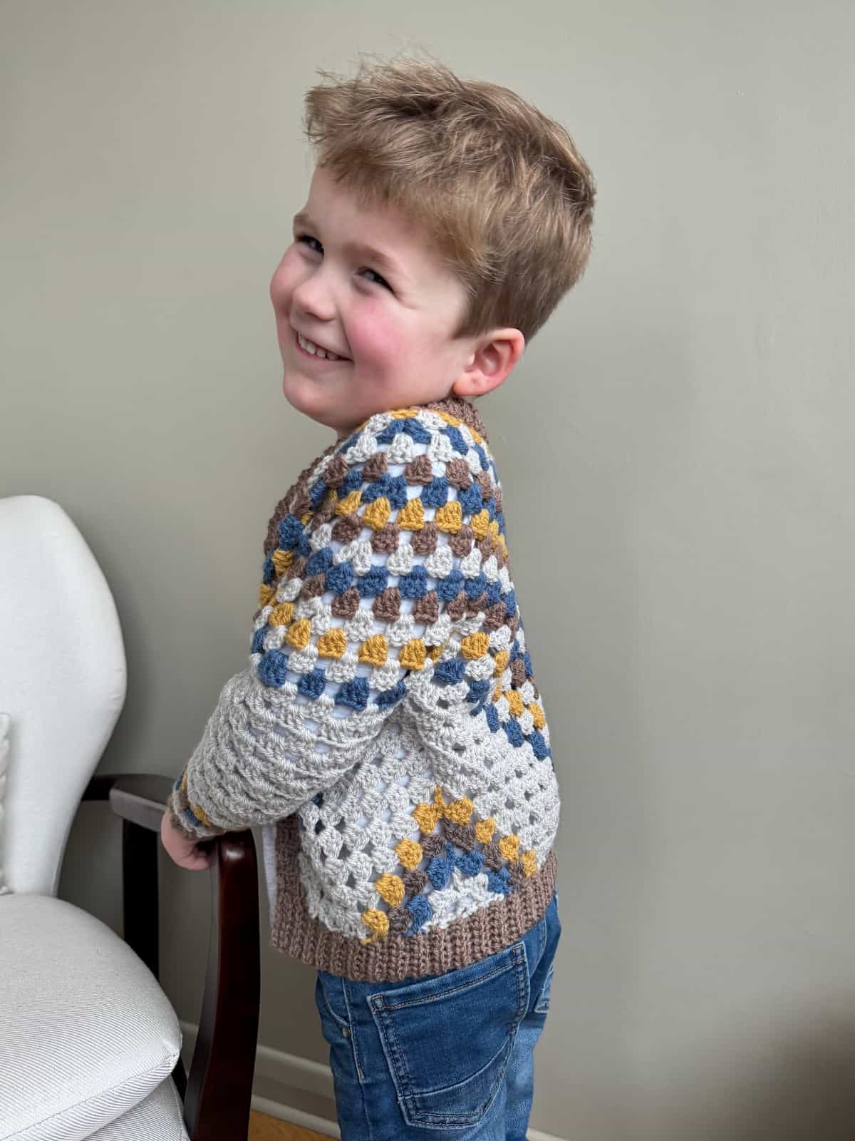 A young boy smiling wearing a crocheted cardigan standing in front of a chair.