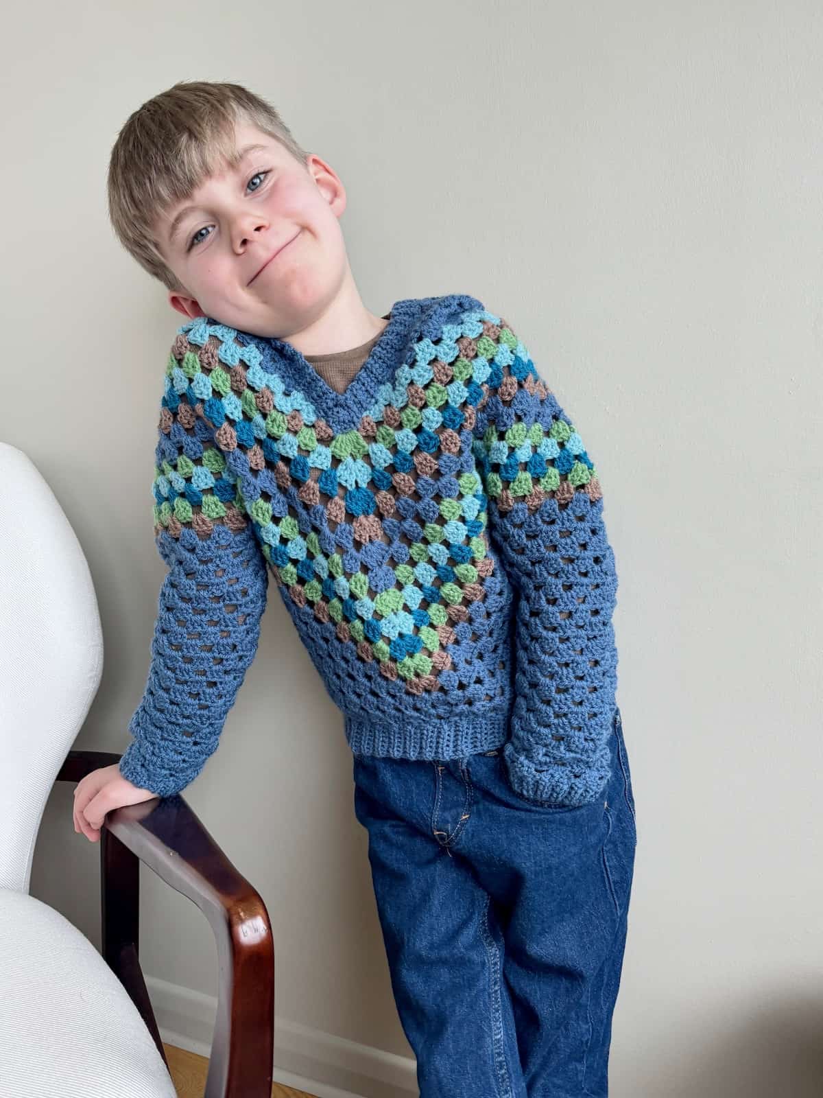 A young boy wearing a crocheted sweater leaning against a chair.