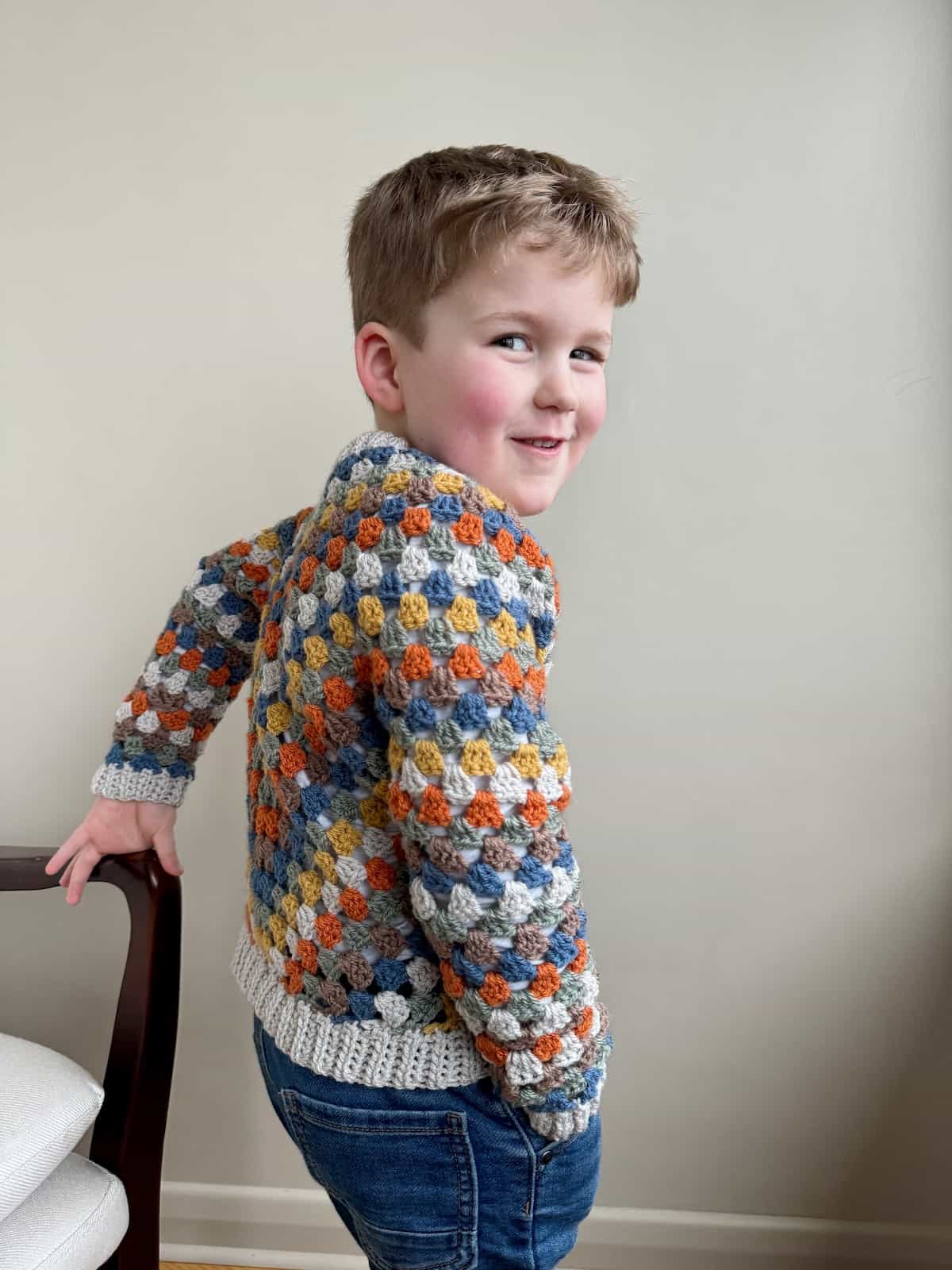 A child in a crocheted sweater poses in front of a chair.