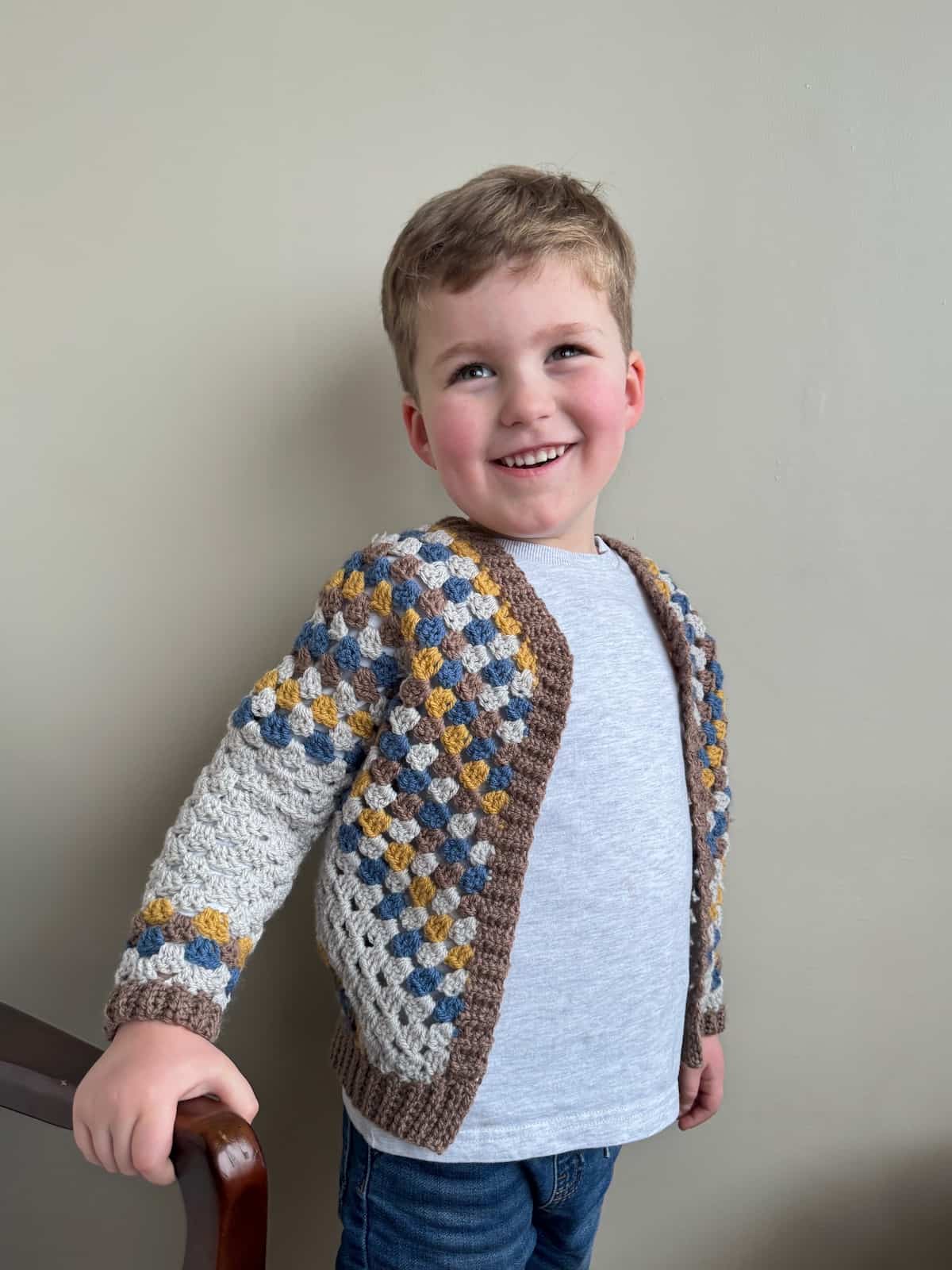A young boy wearing a crocheted cardigan in blue, grey, brown and yellow stripes.