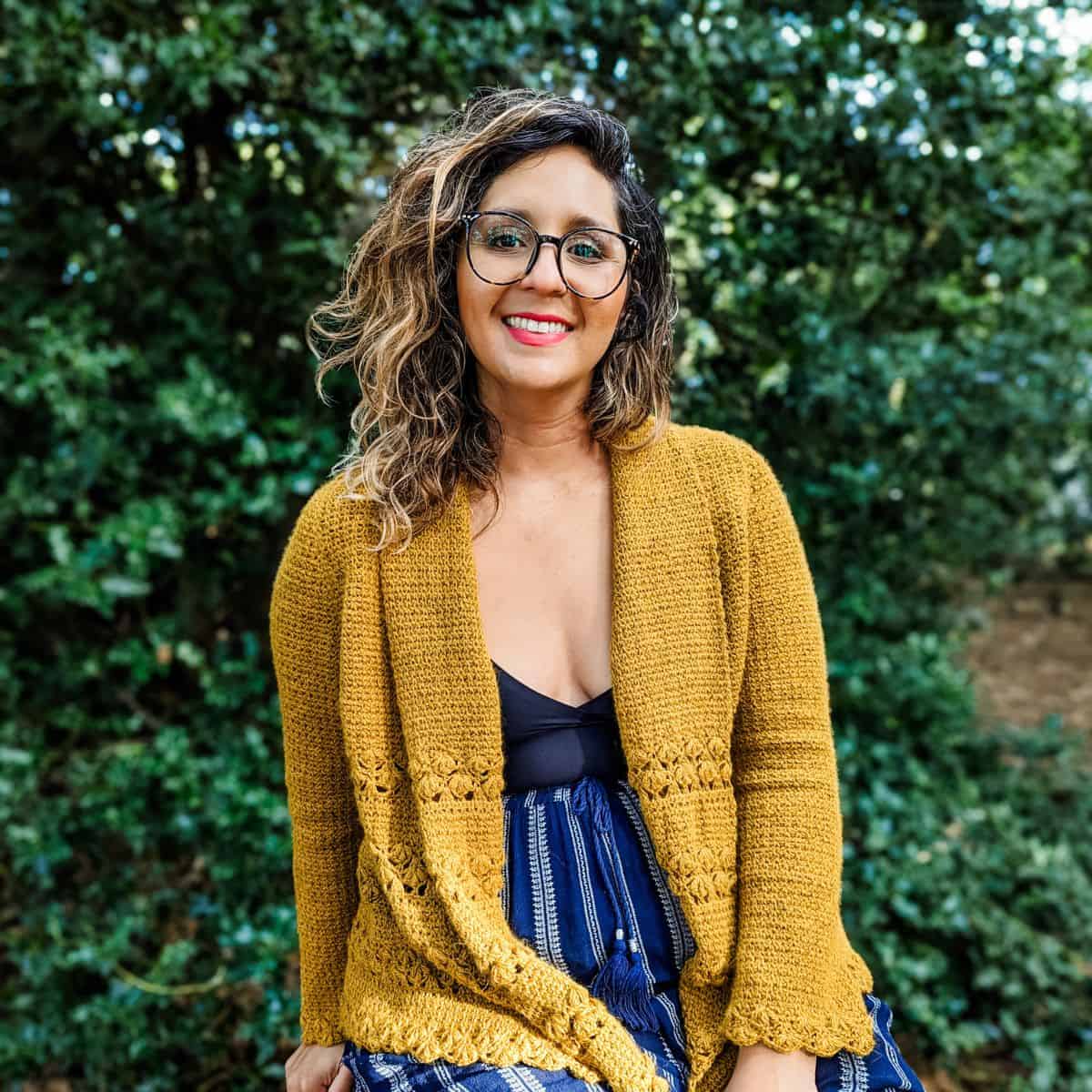 A woman wearing glasses and a yellow crochet cardigan sitting on a bench.