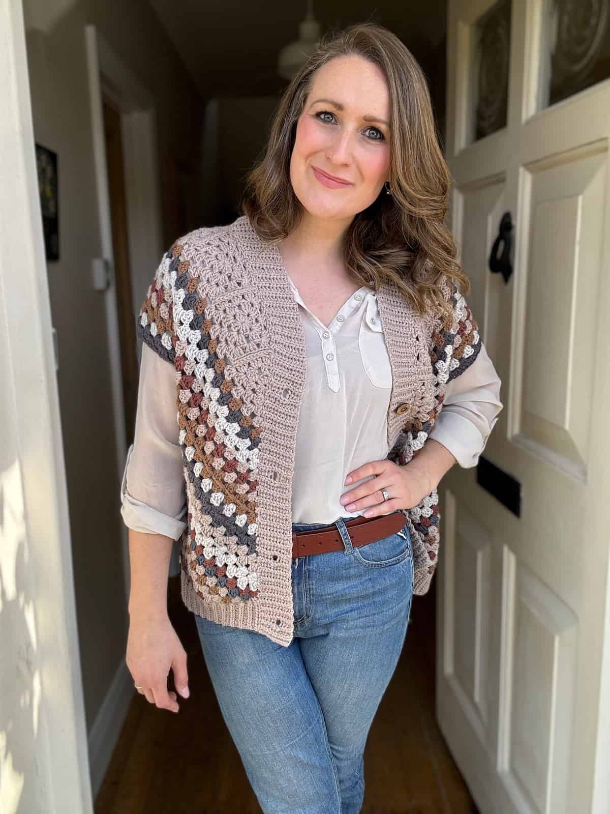 A woman smiling at the camera, standing indoors wearing a granny square crochet jacket over a casual blouse and jeans.