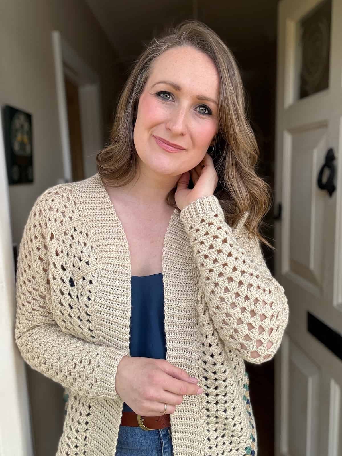 A woman with shoulder-length hair wearing a crochet cardigan poses for the camera indoors.