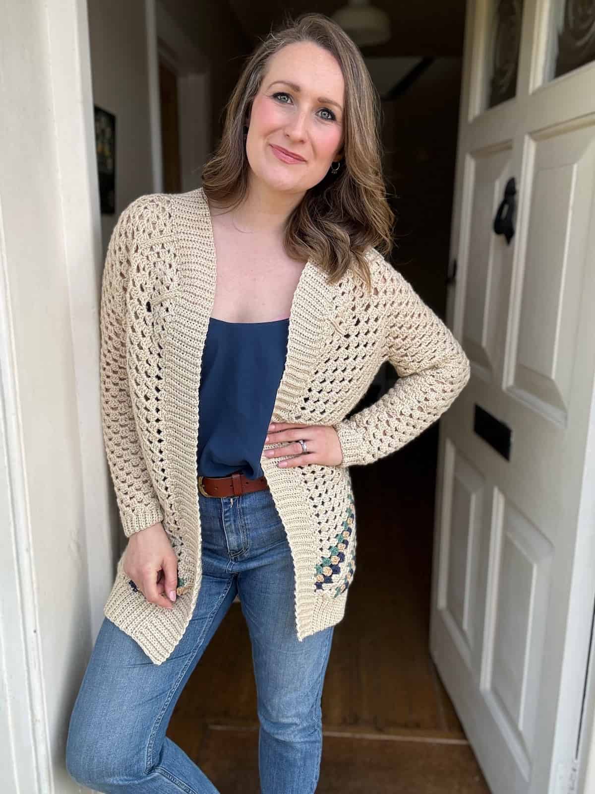 A woman in a beige crochet cardigan and blue top standing in a doorway.