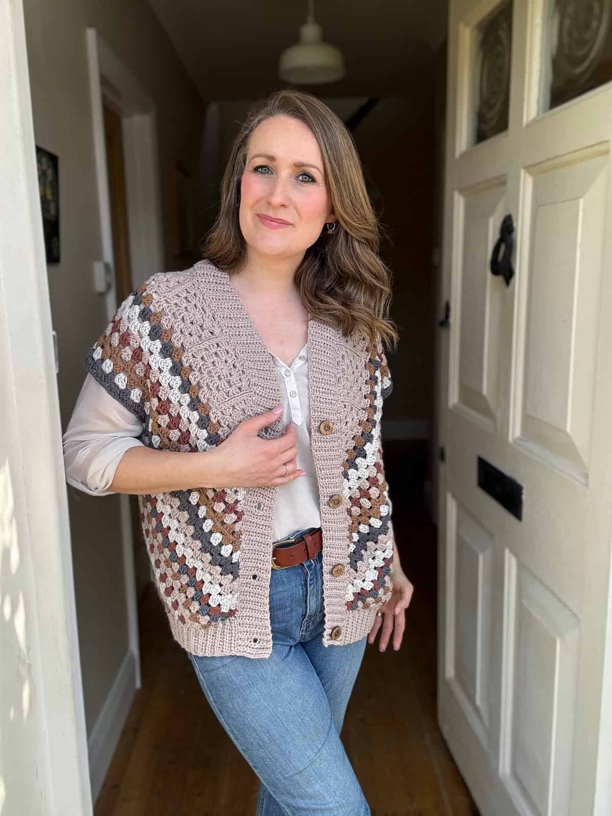 Woman standing in a hallway wearing a short sleeve crochet cardigan over a blouse, paired with jeans.
