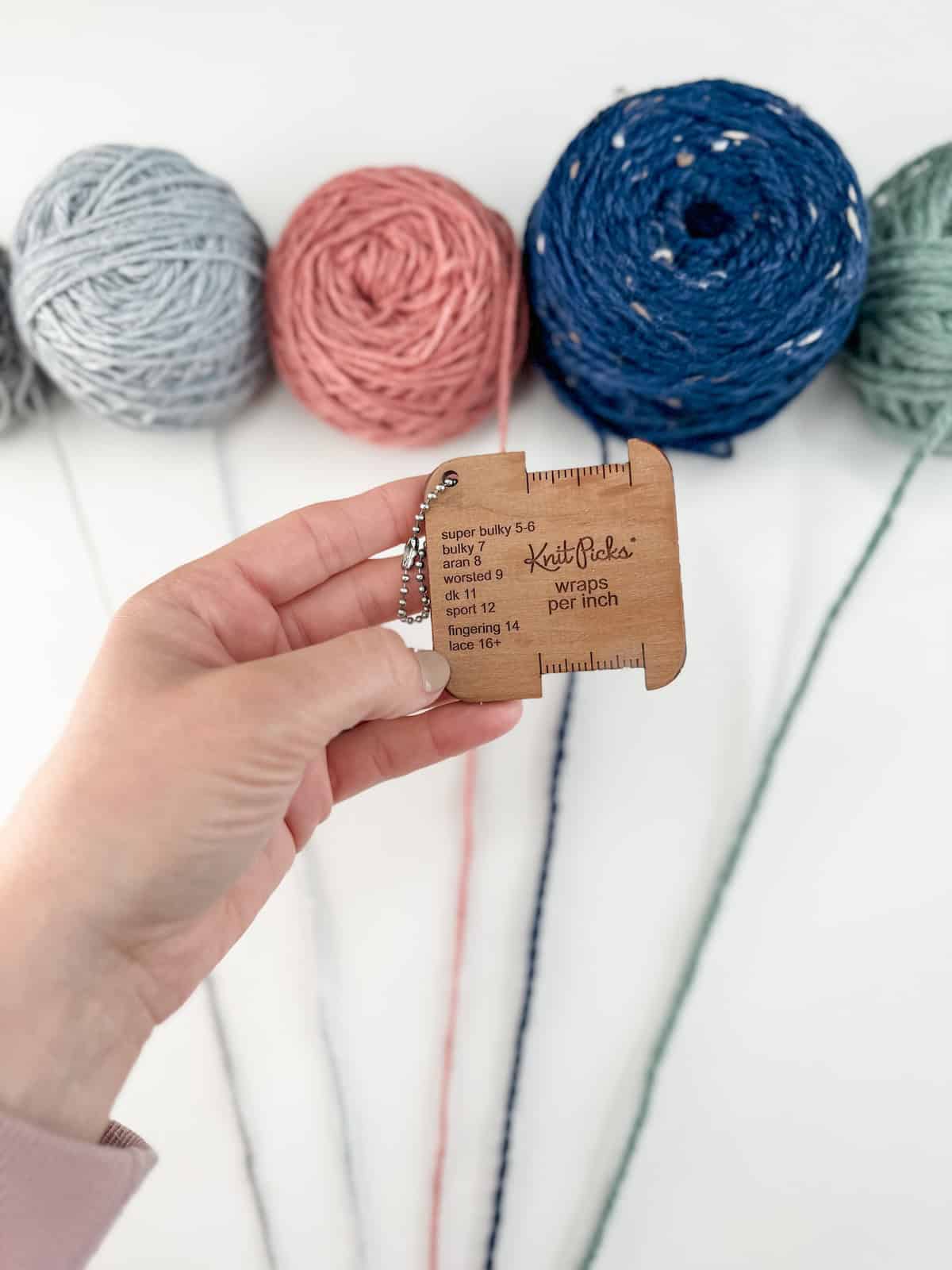 A hand holds a wooden yarn gauge, with four balls of yarn in gray, pink, blue, and green in the background. Yarn strands extend from the balls towards the gauge, illustrating a practical yarn weight chart.