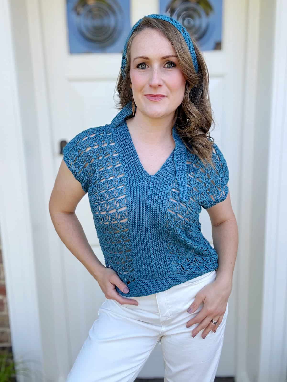 A woman with wavy hair stands in front of a white door, wearing a blue summer crochet top and white pants, with a matching blue headband.