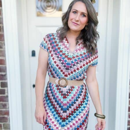 A woman stands in front of a door, wearing a colorful, patterned crochet dress with a beige belt and bracelet.