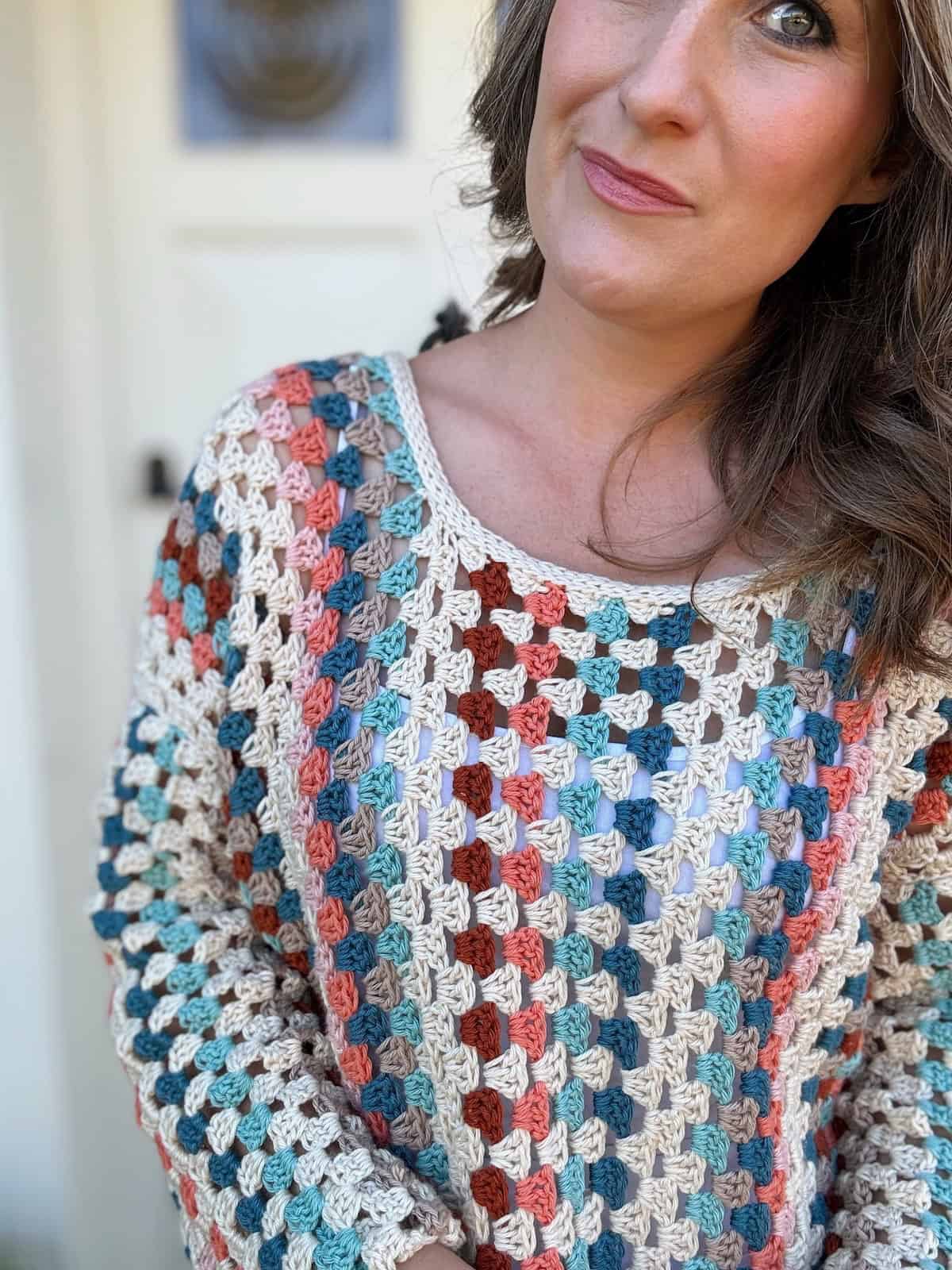 A person with wavy hair wears a colorful crocheted top with a pattern of blue, red, and white squares, standing in front of a white door.