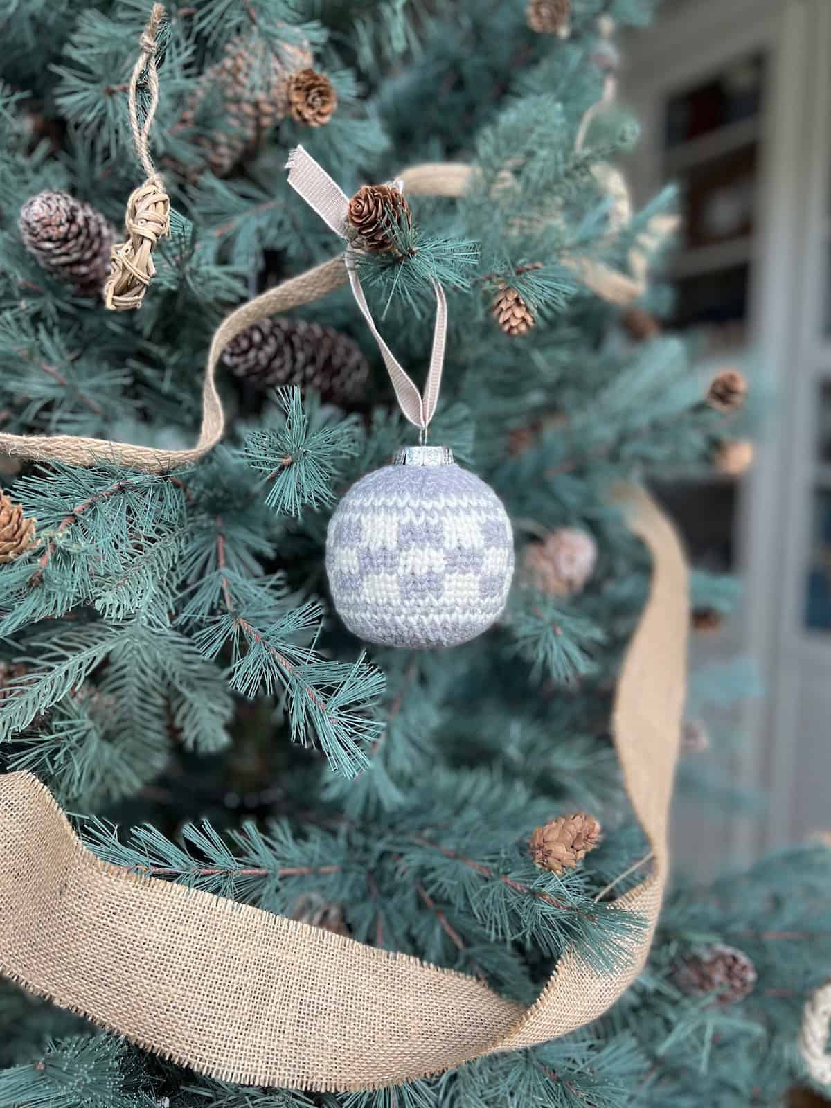 A Christmas tree decorated with a light blue and white gingham patterned crochet ornament, pine cones, and a burlap ribbon.