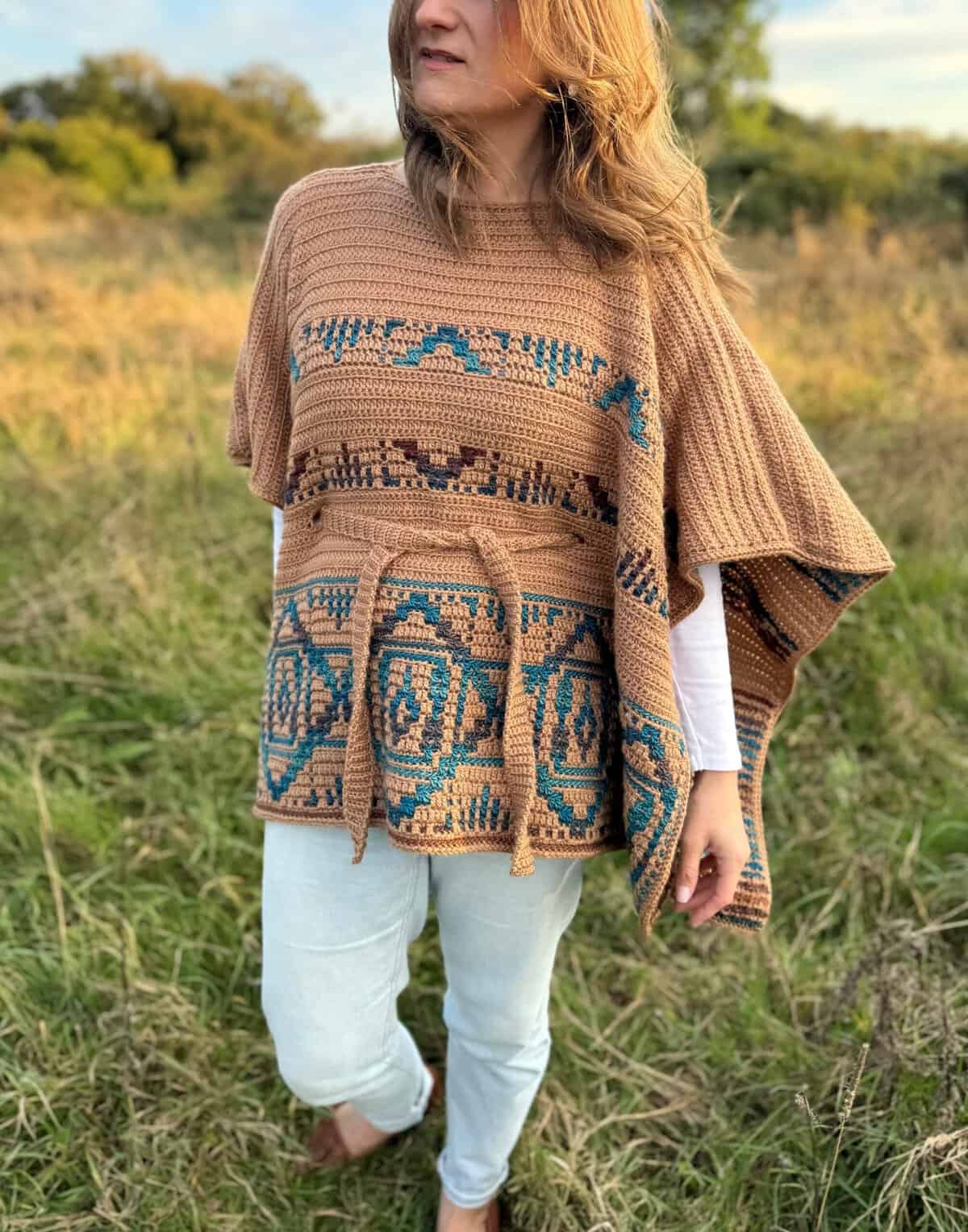 A person stands in a grassy field wearing a patterned poncho and light blue jeans. The poncho is brown with blue and green designs. The background includes trees and a partly cloudy sky.