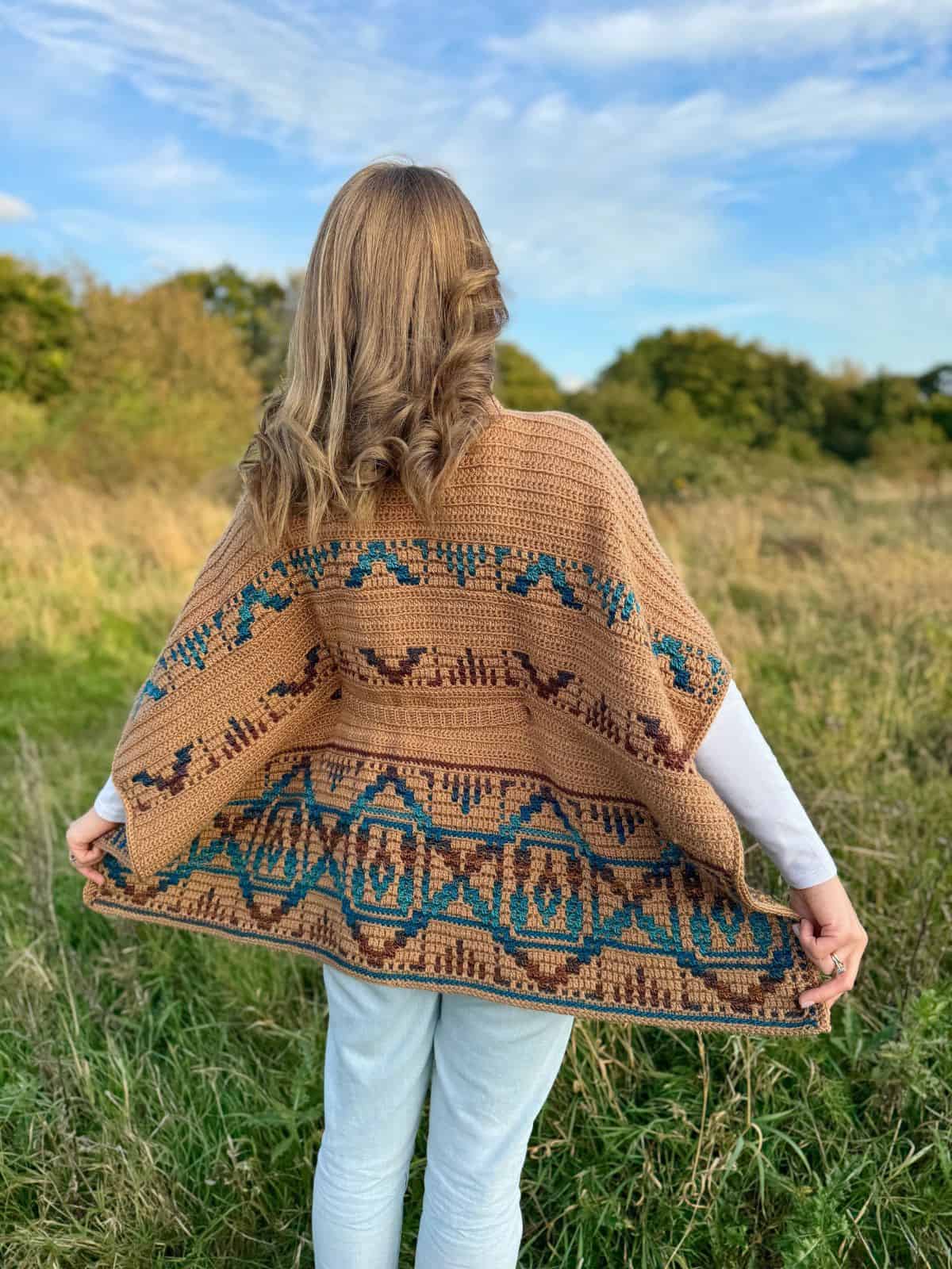 A person stands outdoors in a field with their back to the camera, wrapped in a beautifully crafted crochet poncho featuring intricate geometric patterns.