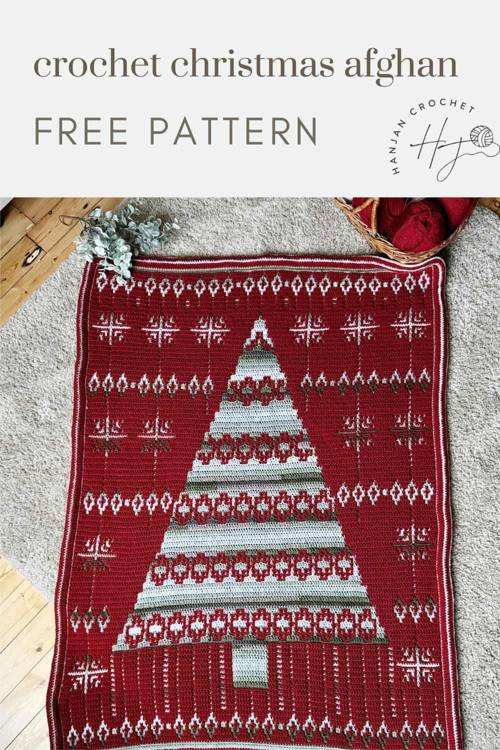 A red crochet Christmas blanket, showcasing a central tree design and geometric patterns, is artfully displayed on a wooden floor with a pine branch nearby.
