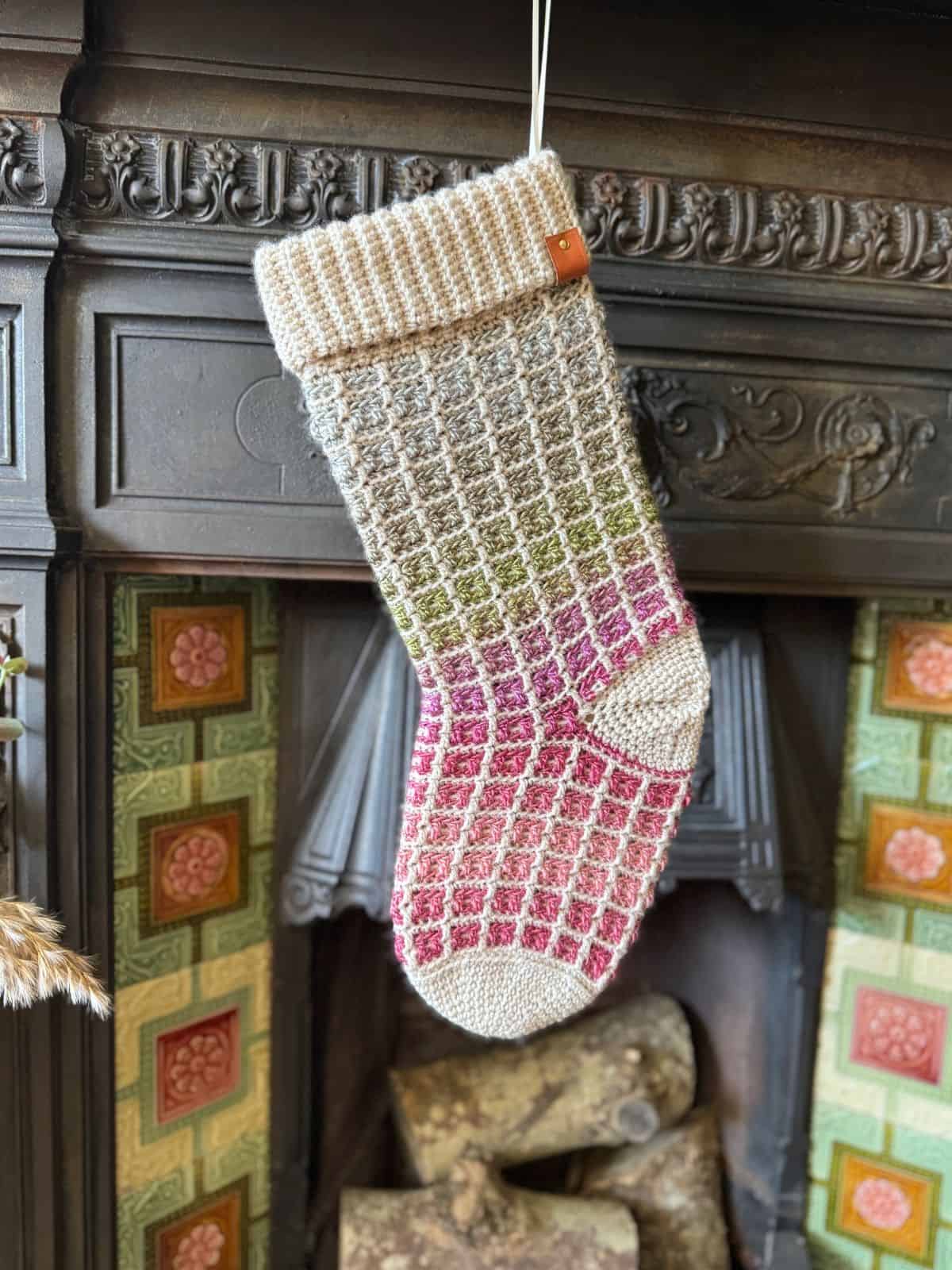 A colorful crocheted Christmas stocking with a geometric pattern hangs on a decorative fireplace, featuring tiles with floral designs.