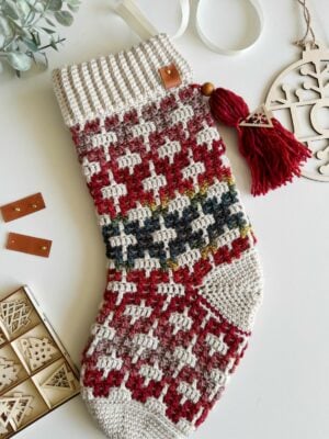 A festive crocheted Christmas stocking featuring a classic red and white pattern, complete with a charming red tassel and accompanied by a wooden ornament. Surrounding the scene are various decorations and craft materials, perfect for those following a crochet Christmas stocking pattern.