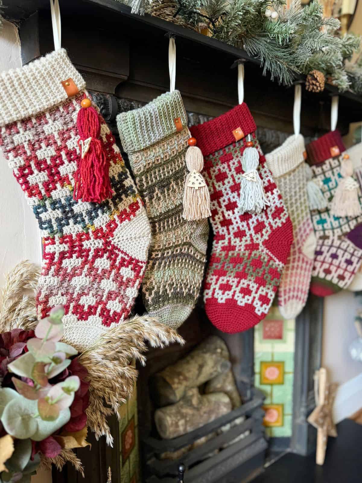 Four knitted Christmas stockings hang on a decorated fireplace, showcasing various winter patterns and colors. Among them, a crochet Christmas stocking pattern adds a unique texture and charm to the festive display.