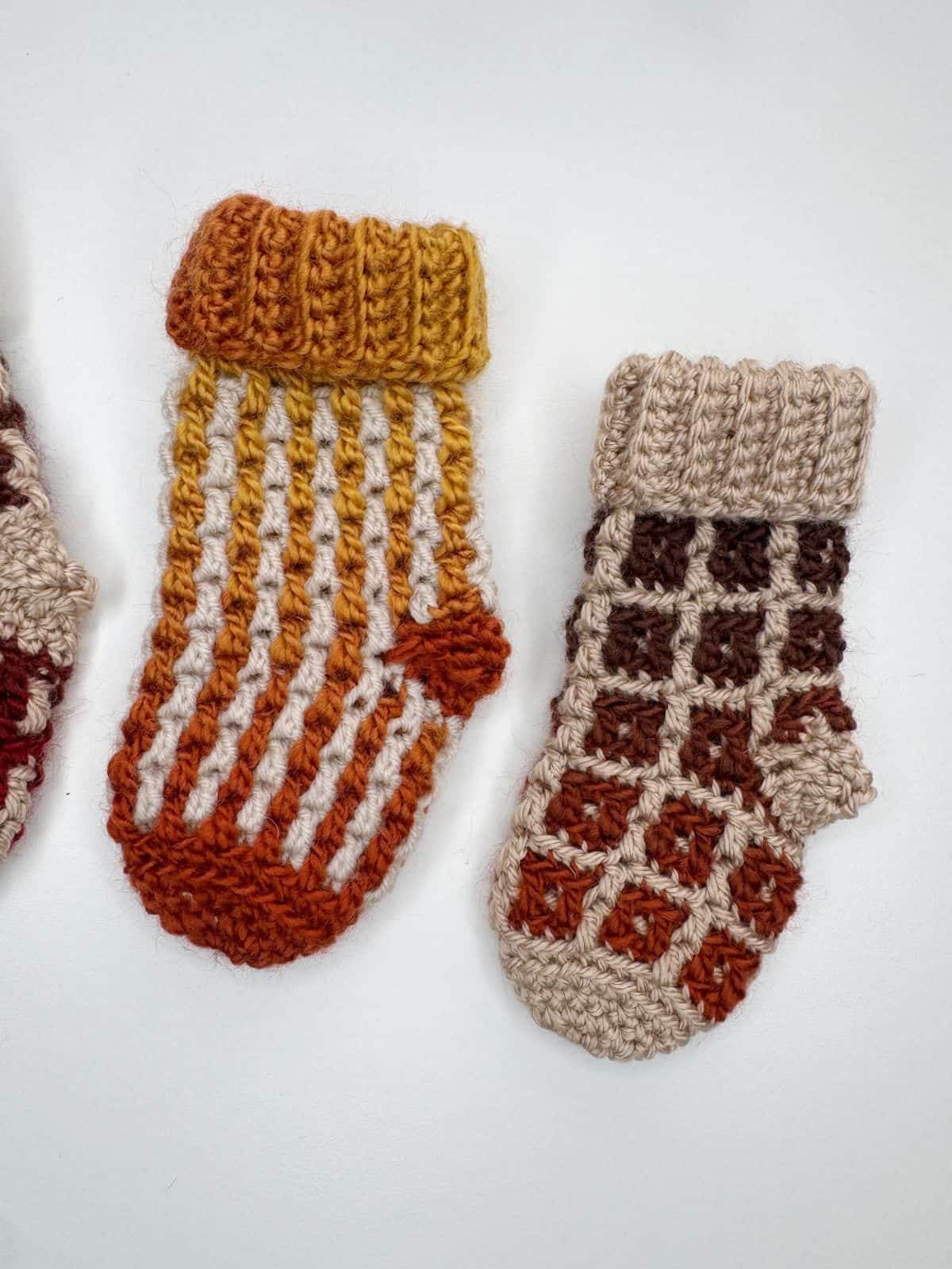Two knitted mittens: one with orange and yellow stripes, and the other with a brown and beige checker pattern, reminiscent of a mini crochet Christmas stocking design, displayed on a white background.