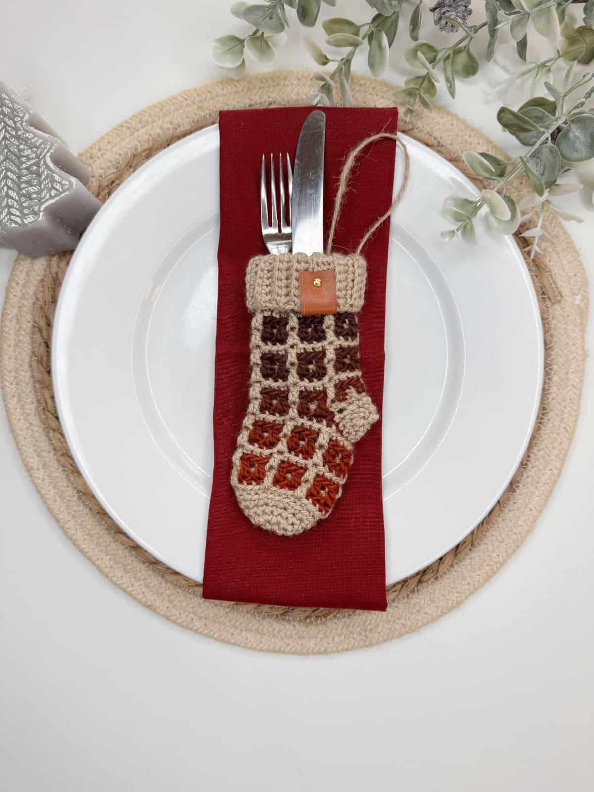 A dining plate with a red napkin and utensils is elegantly decorated with a mini crochet Christmas stocking, resting on a woven placemat accented by greenery.
