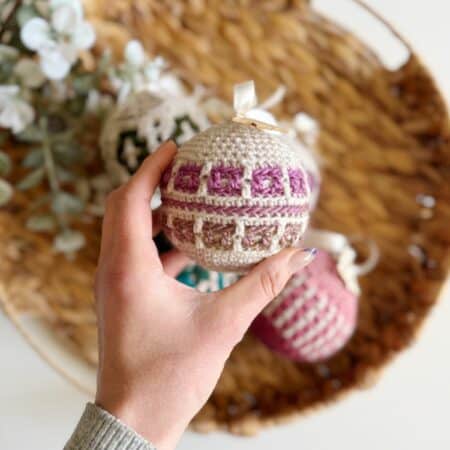 A hand holding a crocheted ornament with a geometric pattern, nestled among crochet tree decorations on a woven tray.