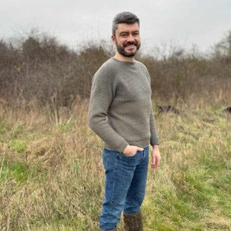 A man stands in a grassy field, wearing a stylish grey crochet sweater, blue jeans, and brown boots, with trees gracefully framing the background.