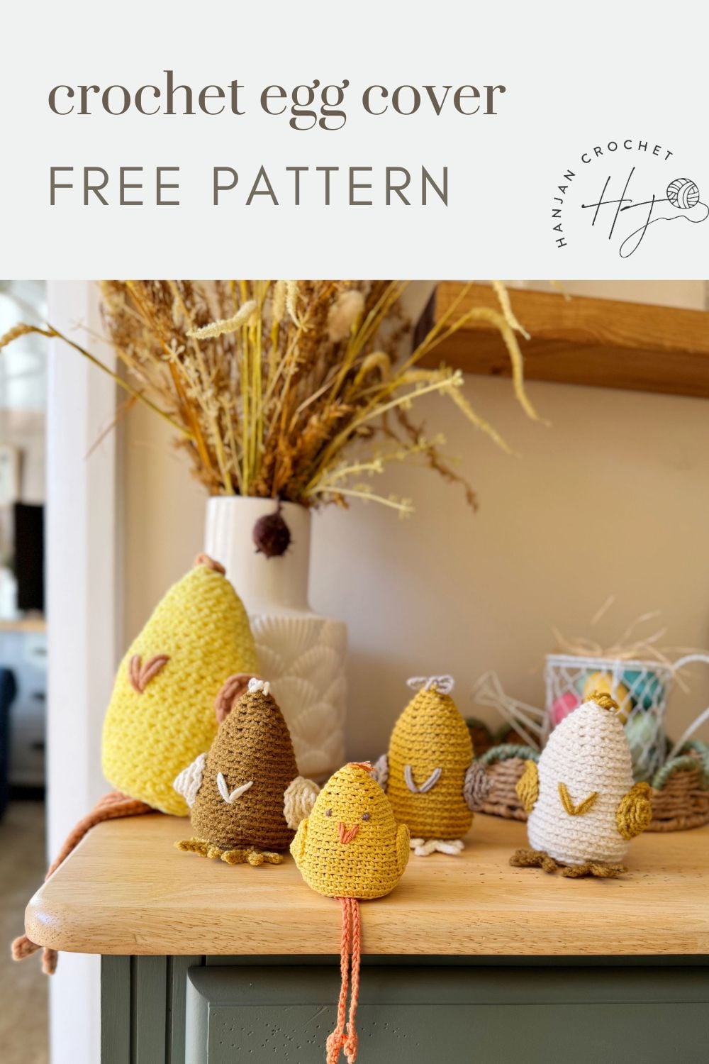 A group of crocheted egg covers shaped like birds in various colors are displayed on a table in front of a vase with dried flowers. Text above announces it as a free crochet chicken pattern.