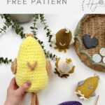 A hand holds a yellow chick egg cozy made from a crochet chicken pattern, surrounded by other crocheted egg cozies and a purple foil-wrapped egg on a white surface.