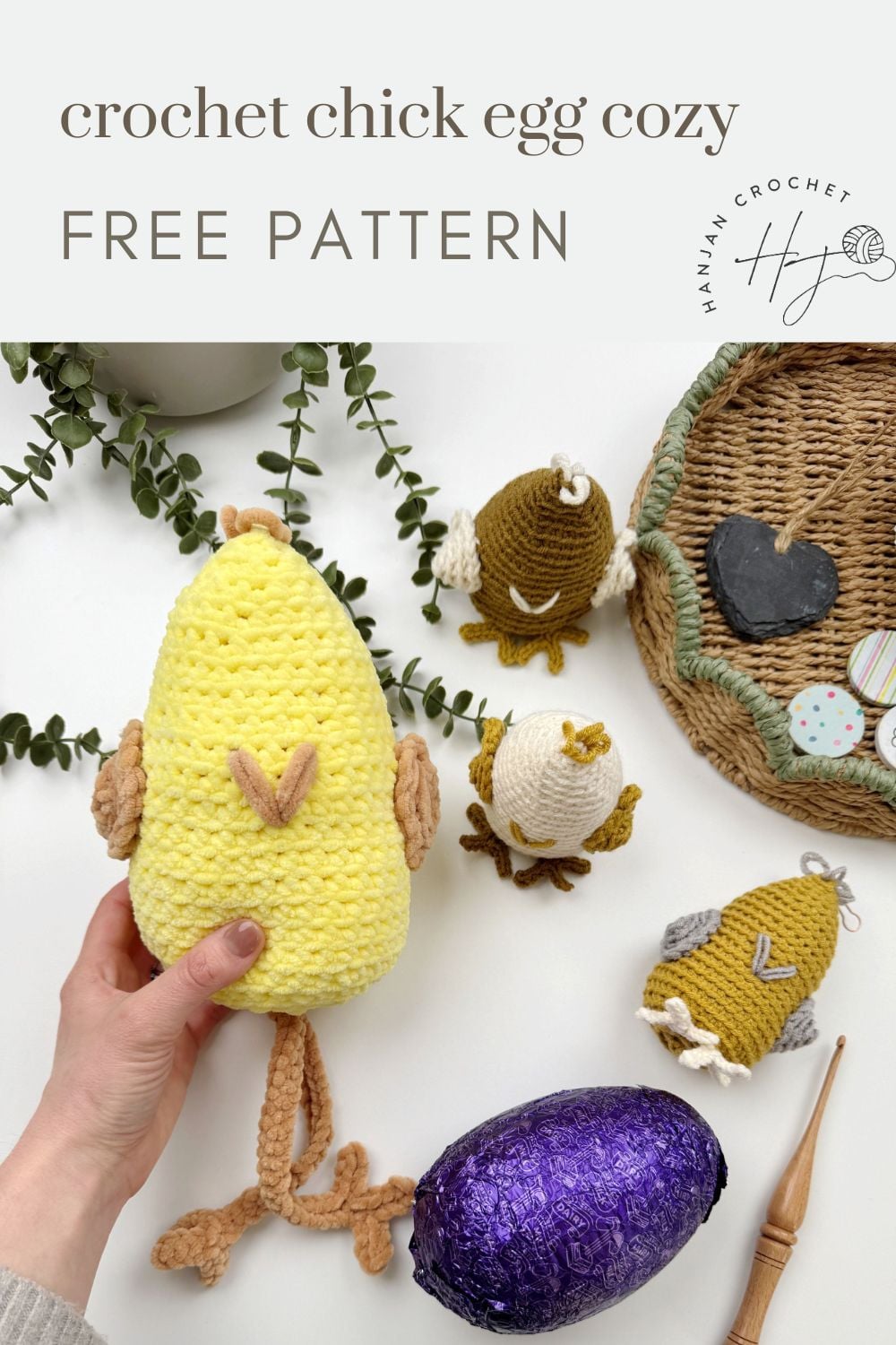 A hand holds a yellow chick egg cozy made from a crochet chicken pattern, surrounded by other crocheted egg cozies and a purple foil-wrapped egg on a white surface.