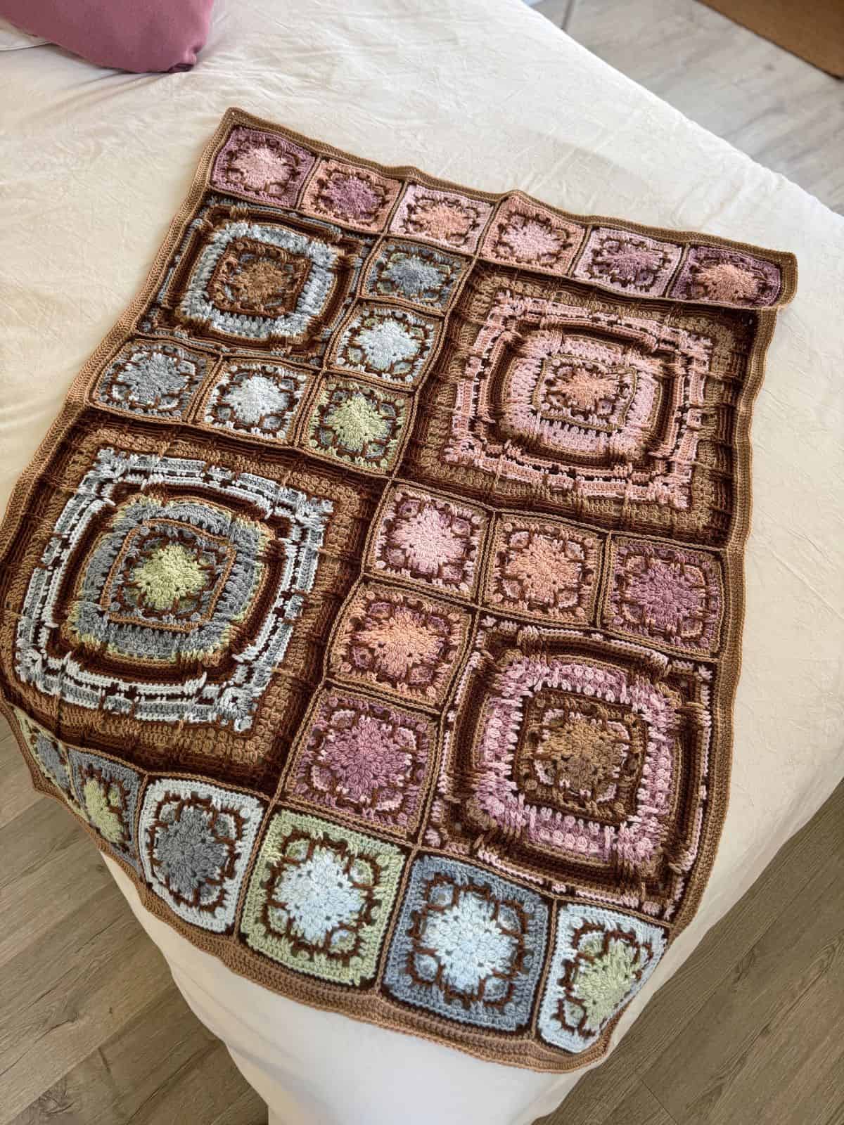 This beautiful crochet blanket, adorned with multicolored square patterns in shades of brown, blue, pink, and green, rests gracefully on a white bed.
