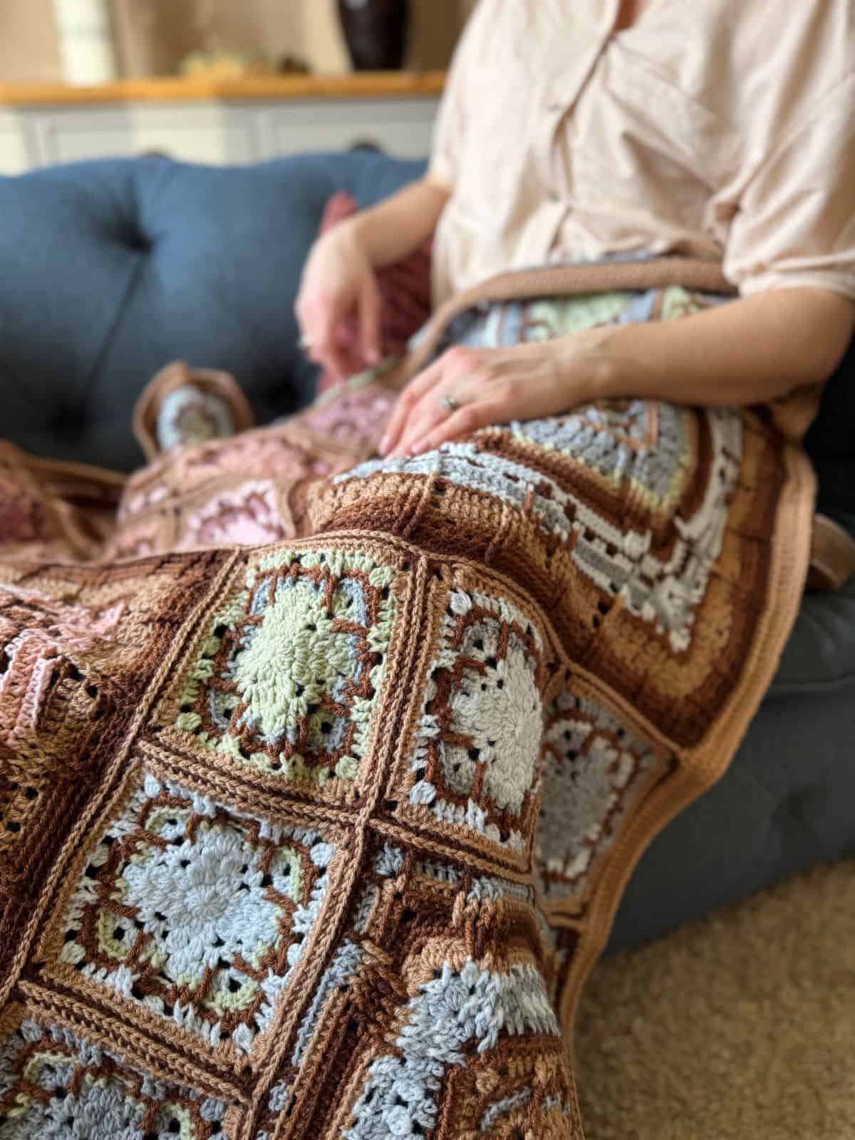 Person sitting on a blue sofa, wrapped in a crocheted quilt with geometric patterns in brown, blue, and white tones.