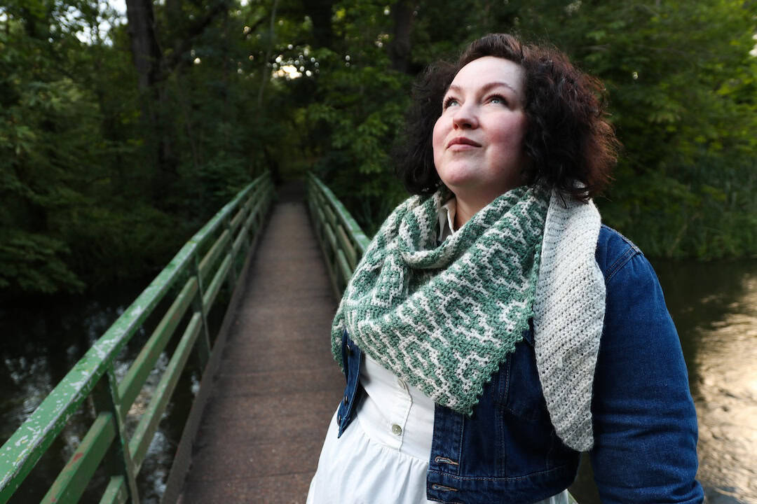 A woman stands on a narrow green bridge in a wooded area, looking upwards and wearing a denim jacket, white shirt, and patterned scarf.