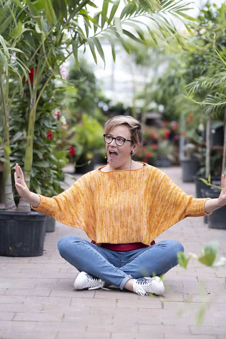 A person wearing glasses and an orange sweater sits cross-legged on a pathway in a greenhouse, with arms extended and mouth open, surrounded by potted plants.