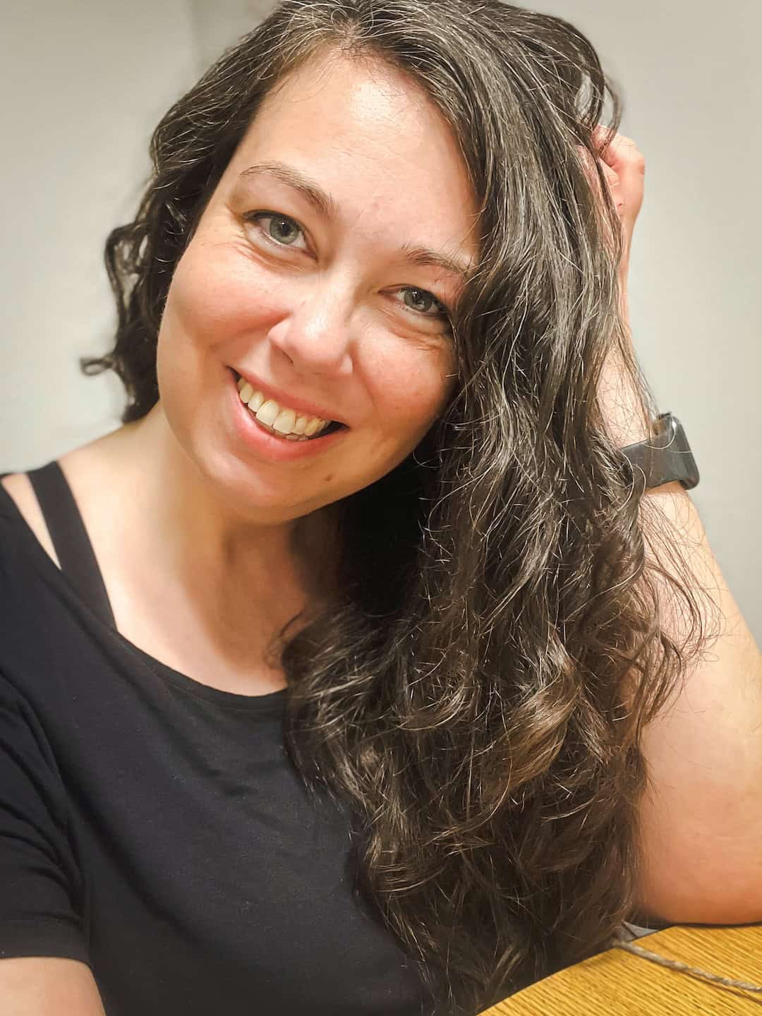 A woman with long wavy brown hair wearing a black shirt and a smartwatch smiles at the camera while sitting at a wooden table.