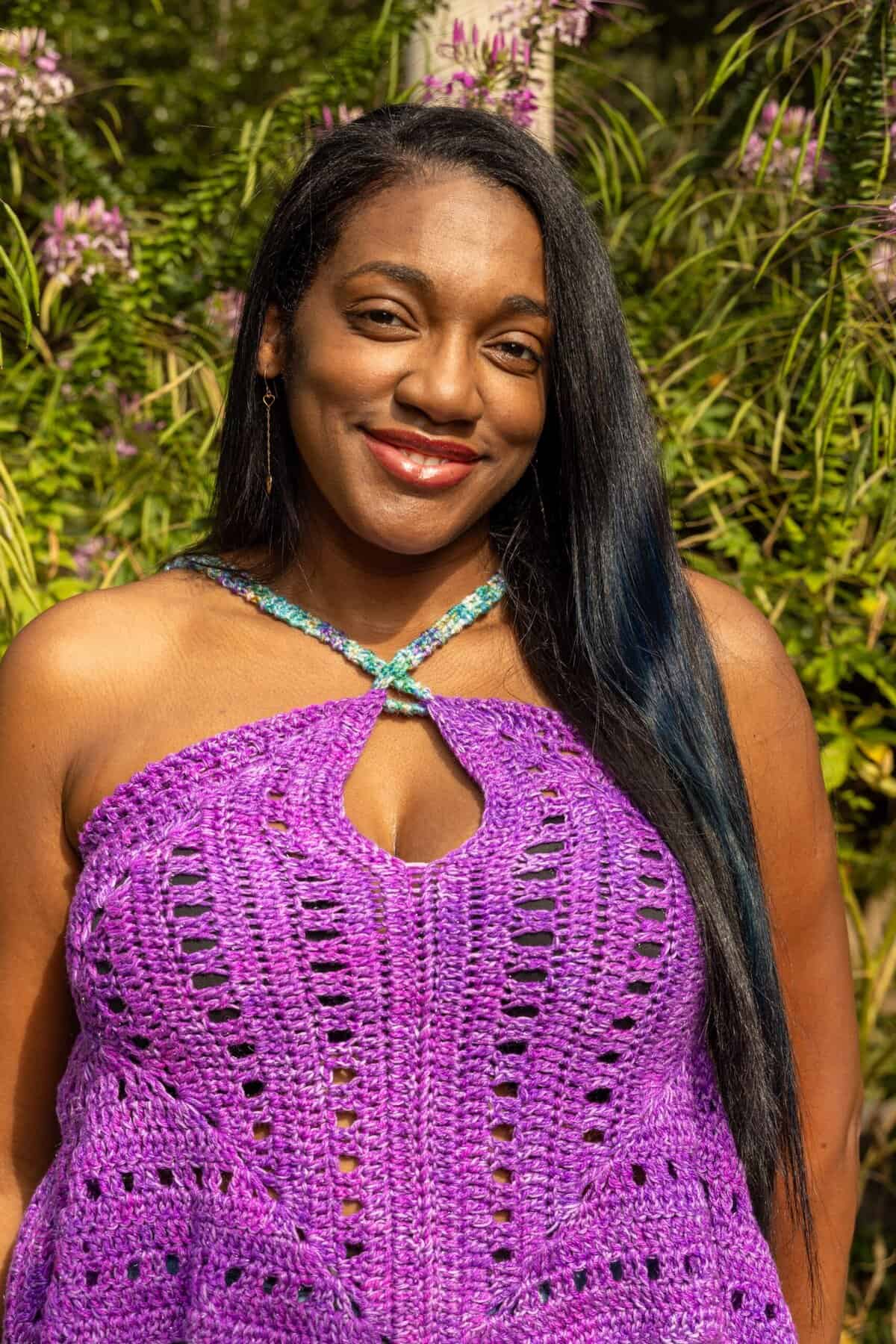 A woman with long black hair wearing a purple crocheted halter top stands outdoors in front of green foliage and purple flowers, smiling at the camera.