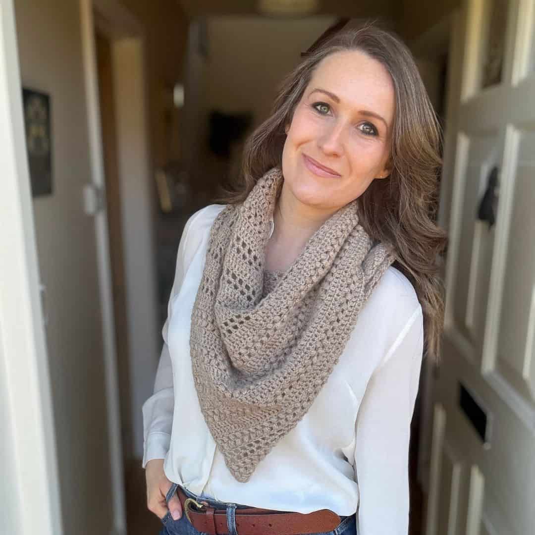 Woman standing indoors, wearing a white blouse, brown belt, and a beige crocheted scarf, with one hand in her pocket, looking at the camera and softly smiling.