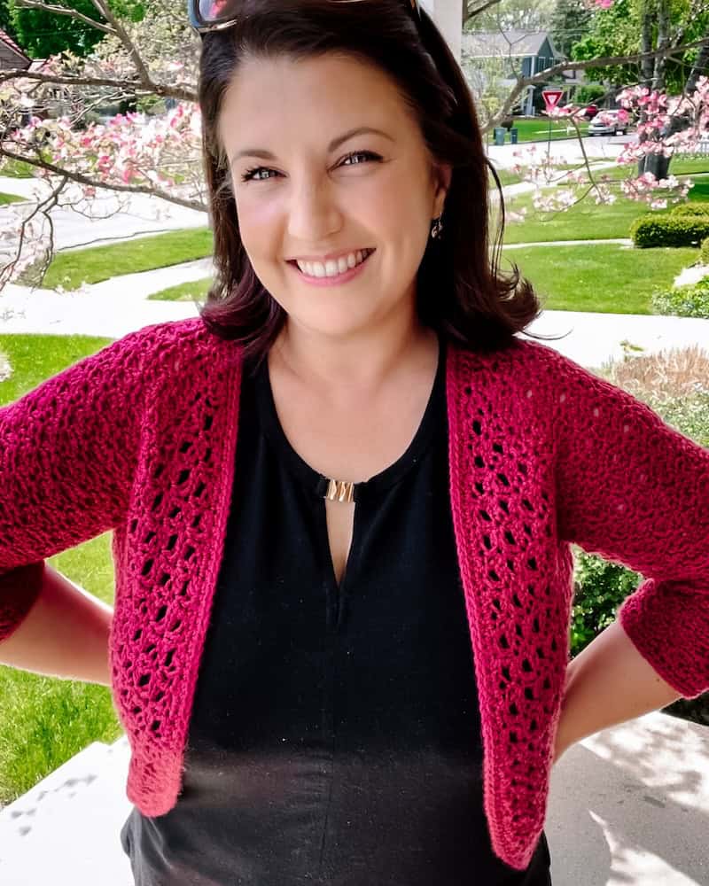 A woman stands outdoors, smiling at the camera. She is wearing a black top and a red crochet cardigan. Spring trees and a green lawn are visible in the background.