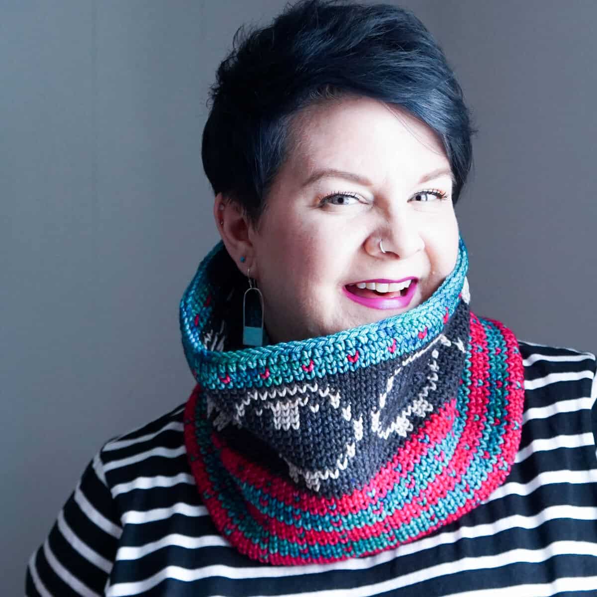A person with short dark hair wears a striped shirt and a colorful knitted cowl, smiling at the camera against a neutral background.