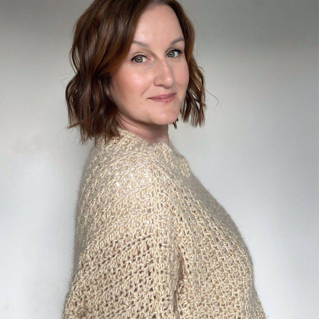 A woman with wavy brown hair, wearing a cream-colored knit sweater, stands against a plain light background and looks at the camera.