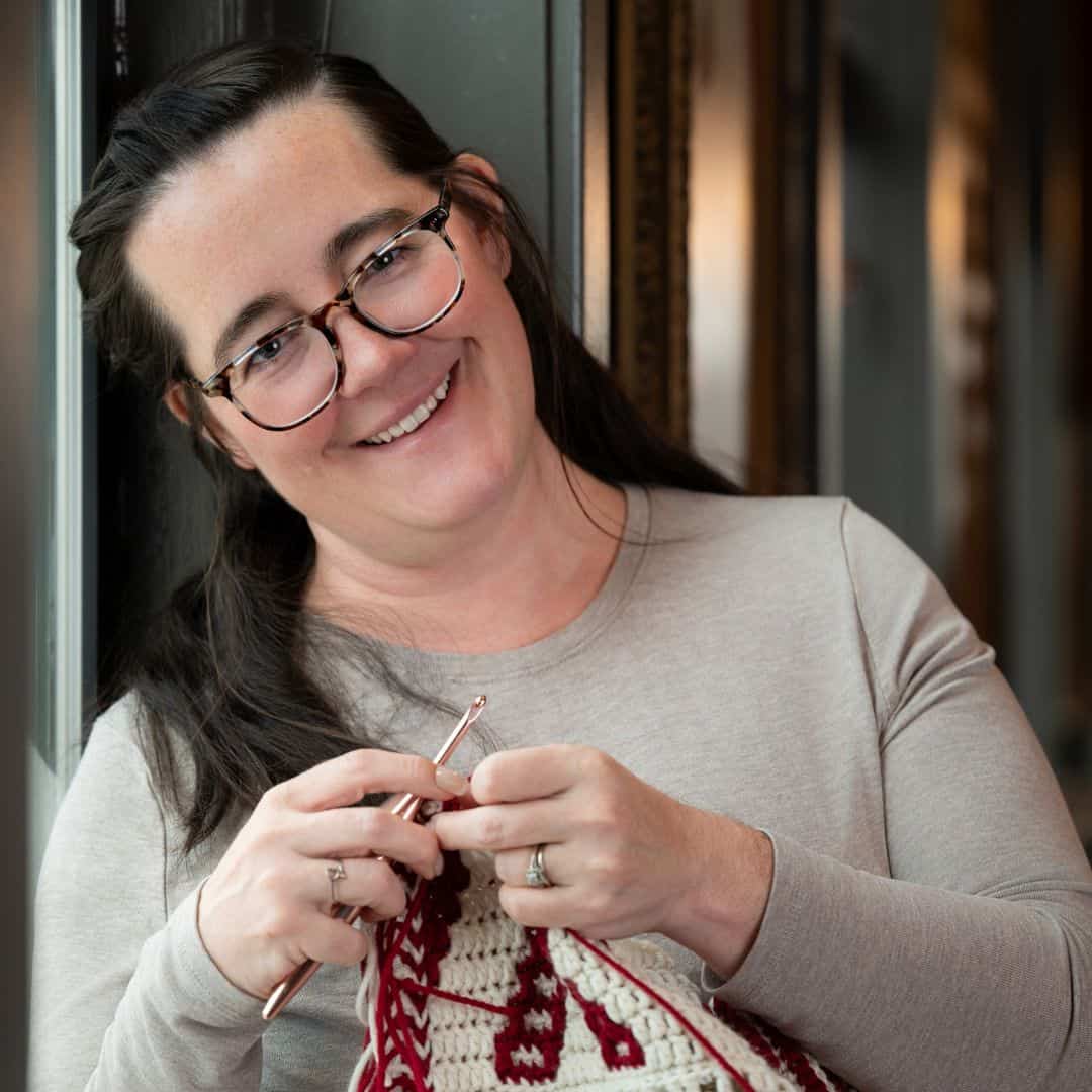 A woman with glasses and long dark hair smiles while crocheting a red and white patterned piece indoors.