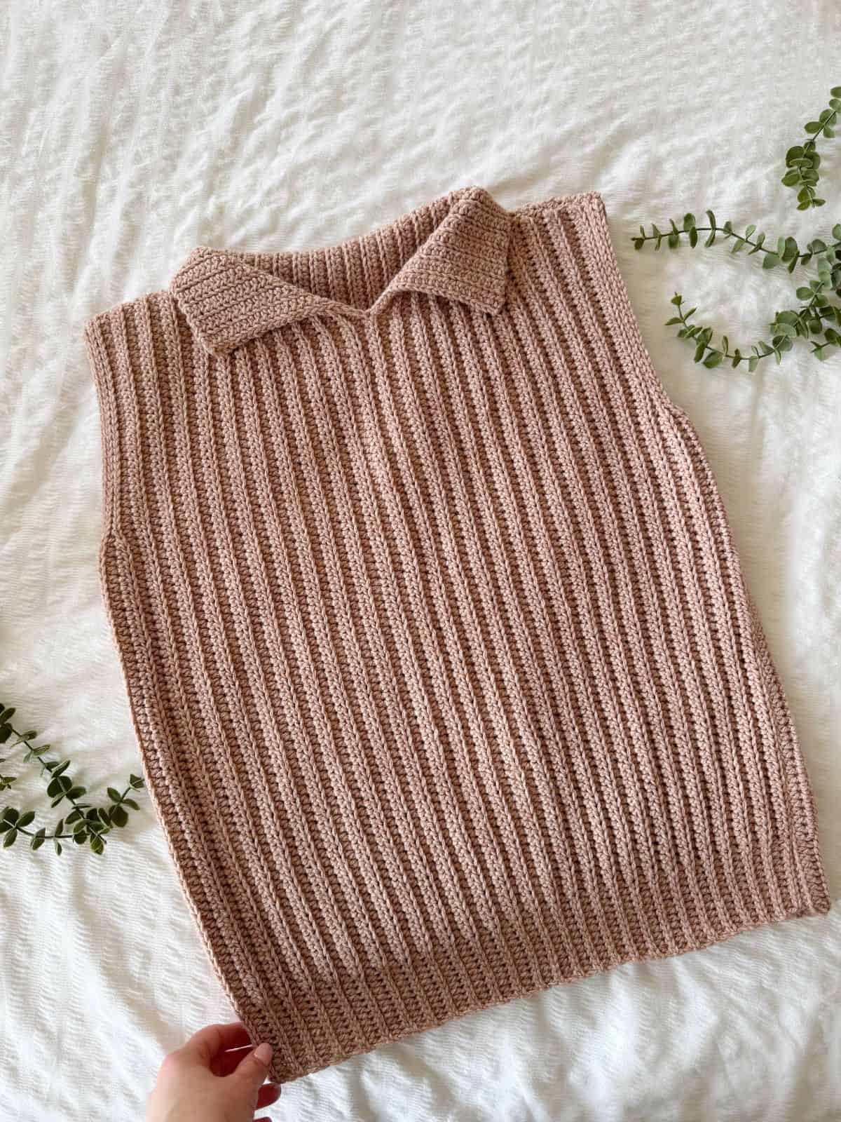 A hand holds the corner of a beige ribbed vest with a collar, laid flat on a white textured surface. Small green plant clippings are arranged nearby, highlighting the modern crochet vest pattern.